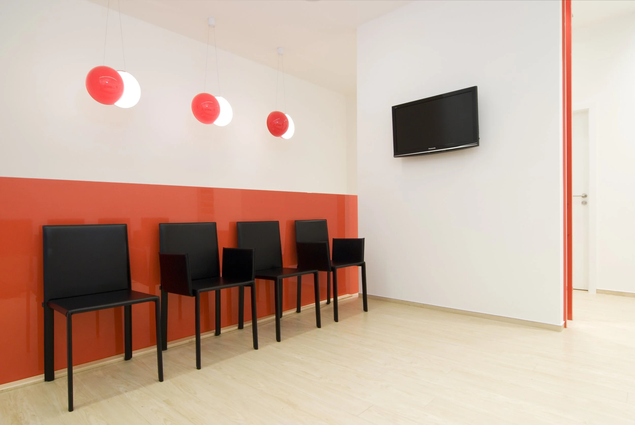 Wartebereich mit fünf schwarzen Stühlen vor einer orangefarbenen Wand, es hängen vier rote Lampen und ein Fernseher an der weißen Wand