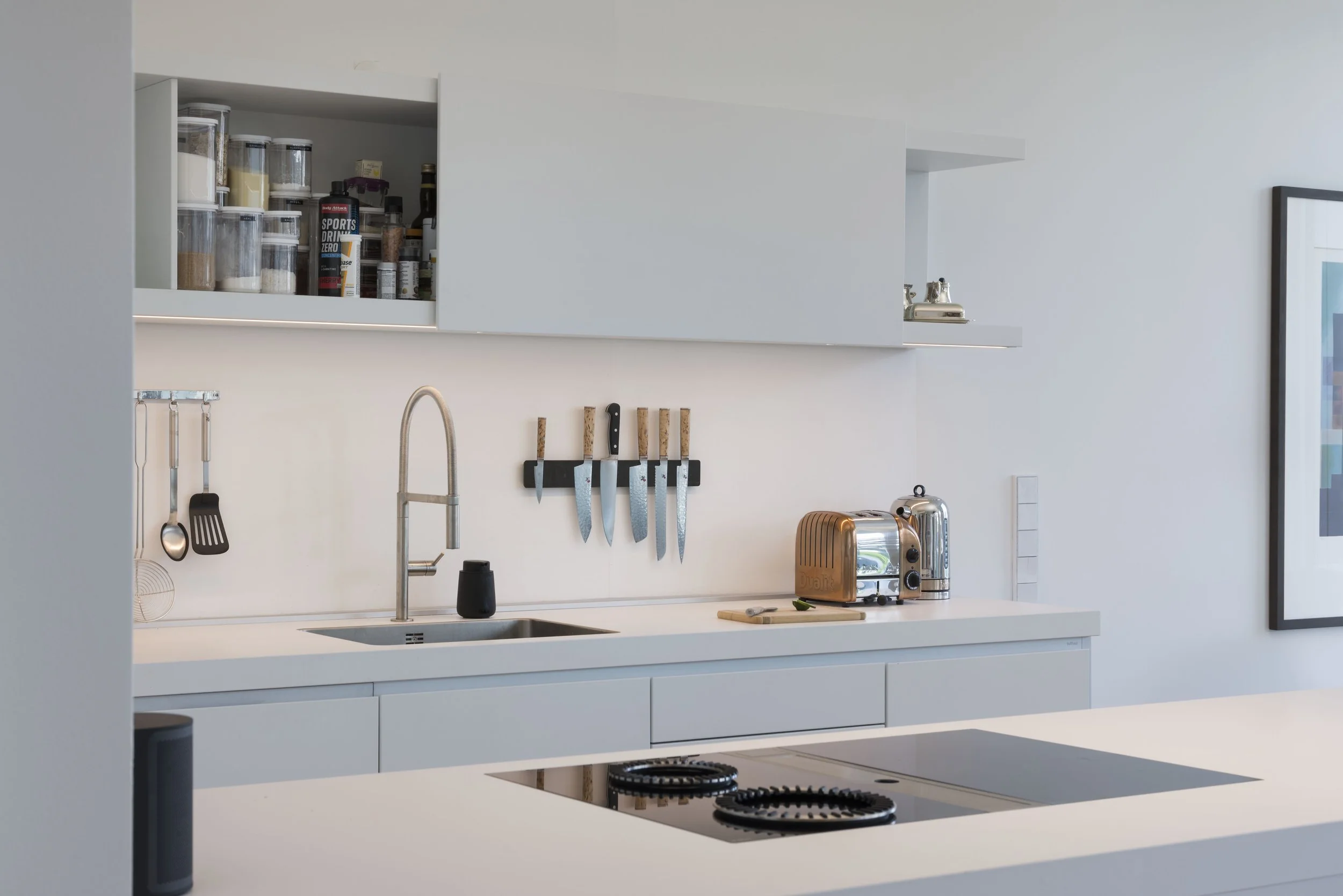 Moderne, minimalistische Küche mit weißen Schränken, Herd auf der Insel, Spüle, Messerblock, Toaster, Wasserkocher, Wandhaken mit Kochutensilien, offene obere Schränke mit Vorräten, Rahmenkunst an der Wand