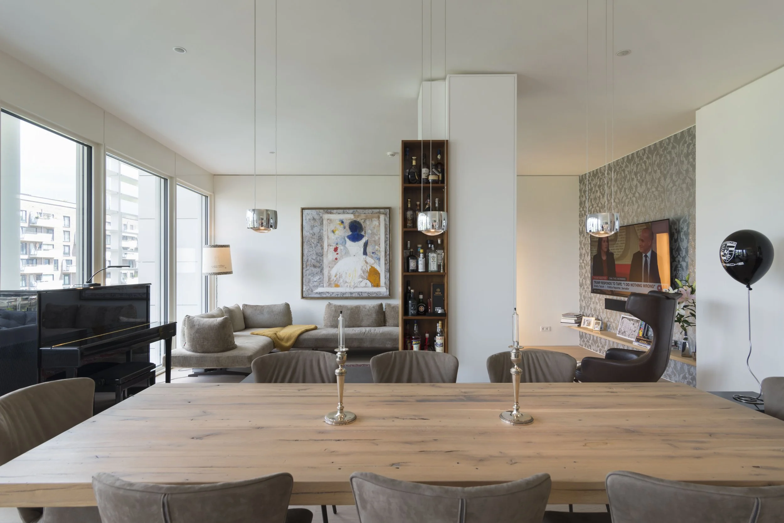 Modernes, helles Wohnzimmer mit offener Gestaltung, großes Holztisch mit Stühlen, Sofa, Fernseher, Kunstwerke, Stehlampen, dekorative Gegenstände, große Fenster mit Blick auf Stadt