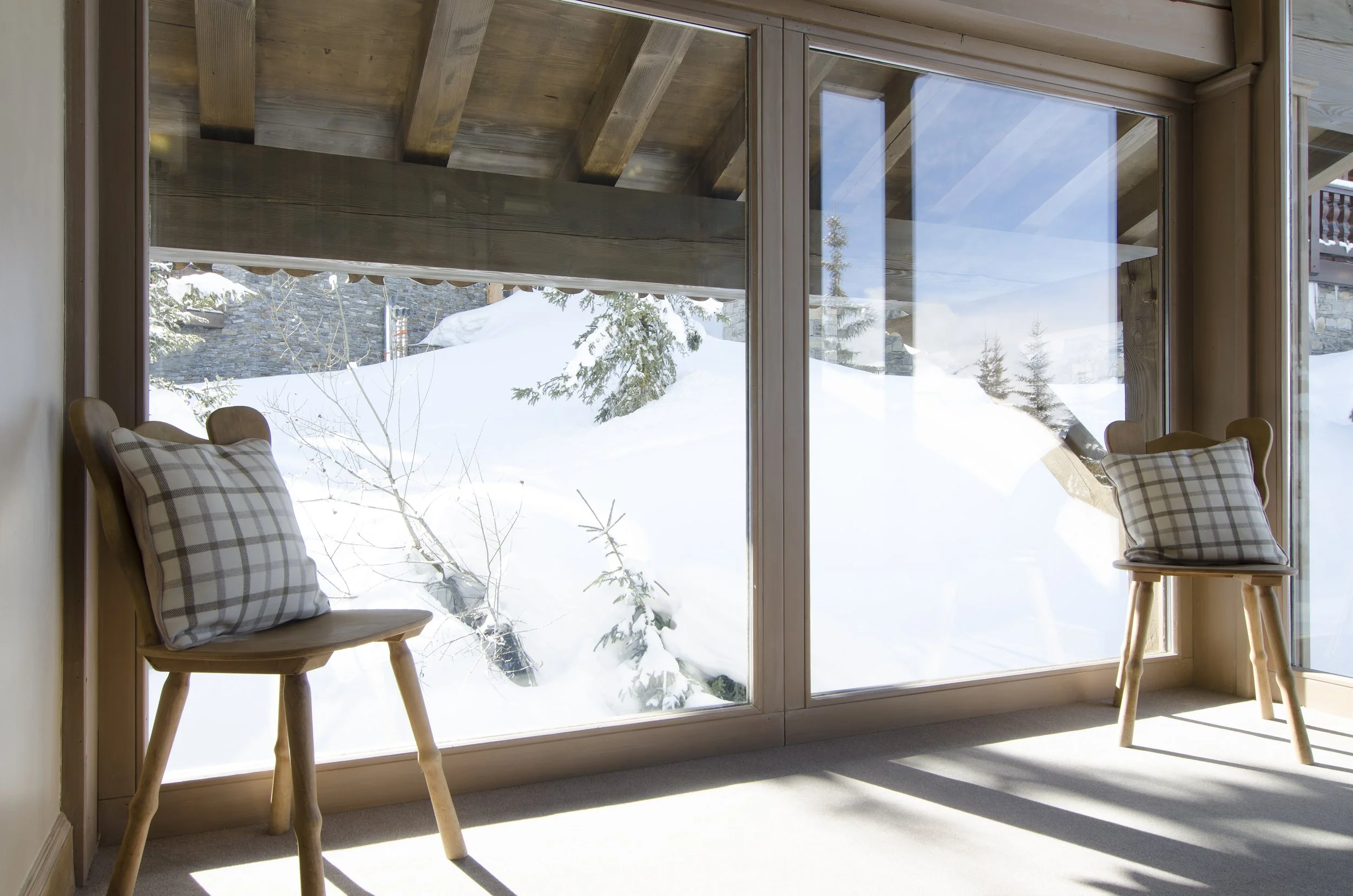 Blick durch große Fenster in eine schneebedeckte Berglandschaft mit Kiefern und einem Steinhaufen.