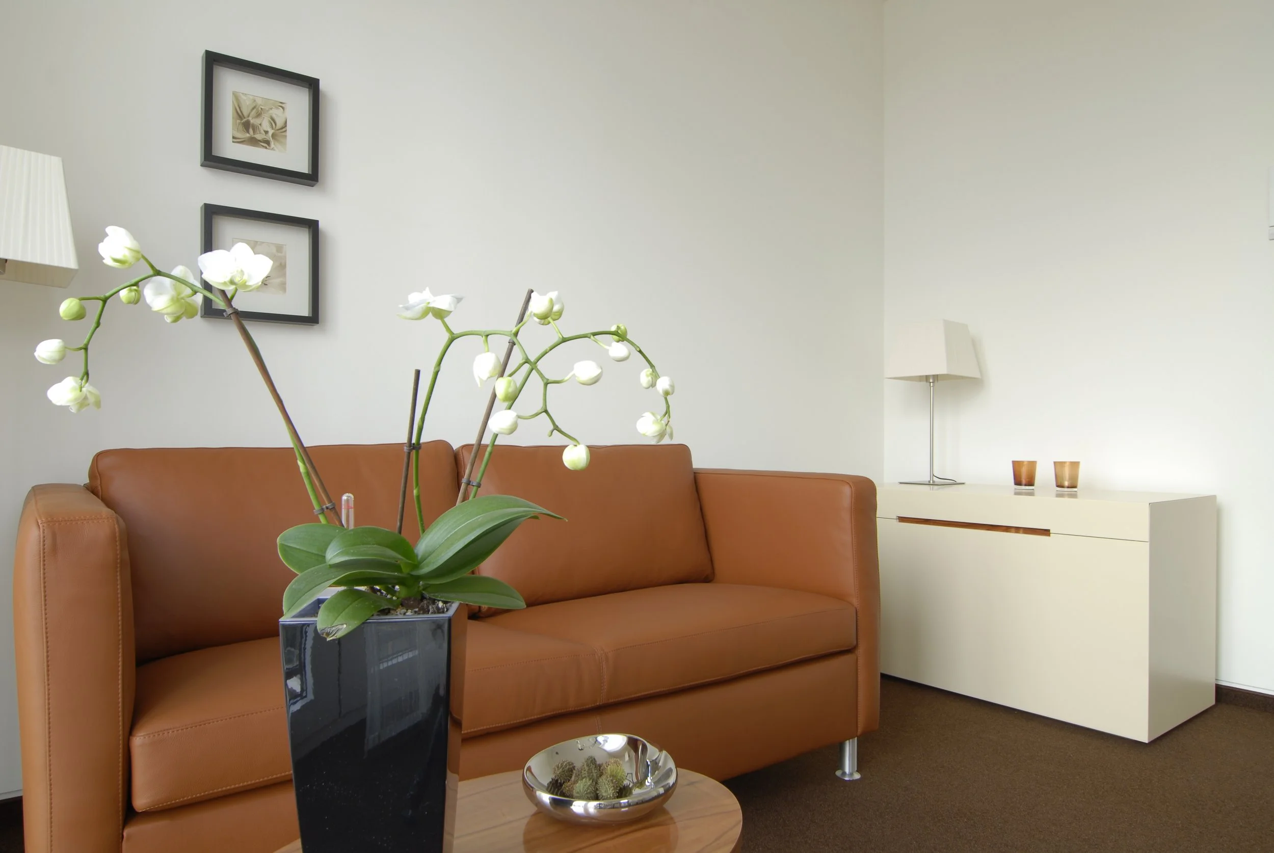 Ein Wohnzimmer mit einem braunen Ledersofa, einem weißen Sideboard, zwei kleinen Kerzen und einem weißen Lampenschirm. Es gibt eine Orchidee in einer schwarzen Vase auf einem runden Holztablett und zwei Bilder auf der Wand.
