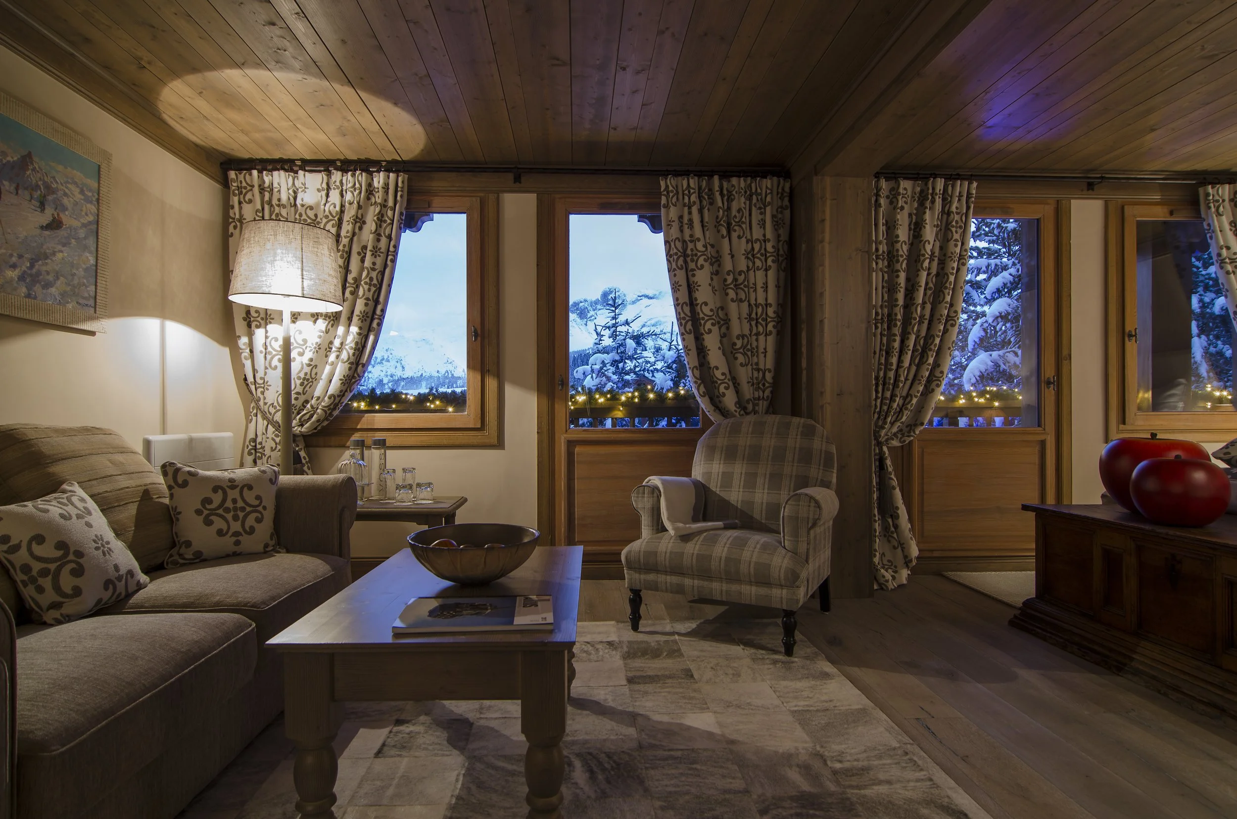 Gemütliches Wohnzimmer mit Holzboden, braunem Sofa, beiger Armchair, Stehlampe, mit Blick auf verschneite Berge durch mehrere Fenster, festlich dekorierte Fensterbank mit Lichterketten, dekorative Vasen auf Sideboard.