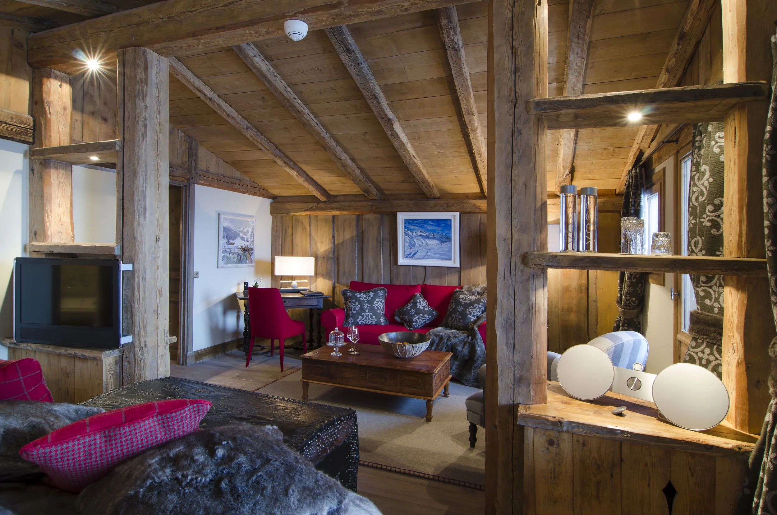 Gemütliches Wohnzimmer mit Holzwänden und -Decken, rotem Sofa und mehreren dekorativen Kissen, einem Holztisch, einem kleinen Schreibtisch mit Lampe, einem Fernseher und einem Fenster mit Scheuklappen.