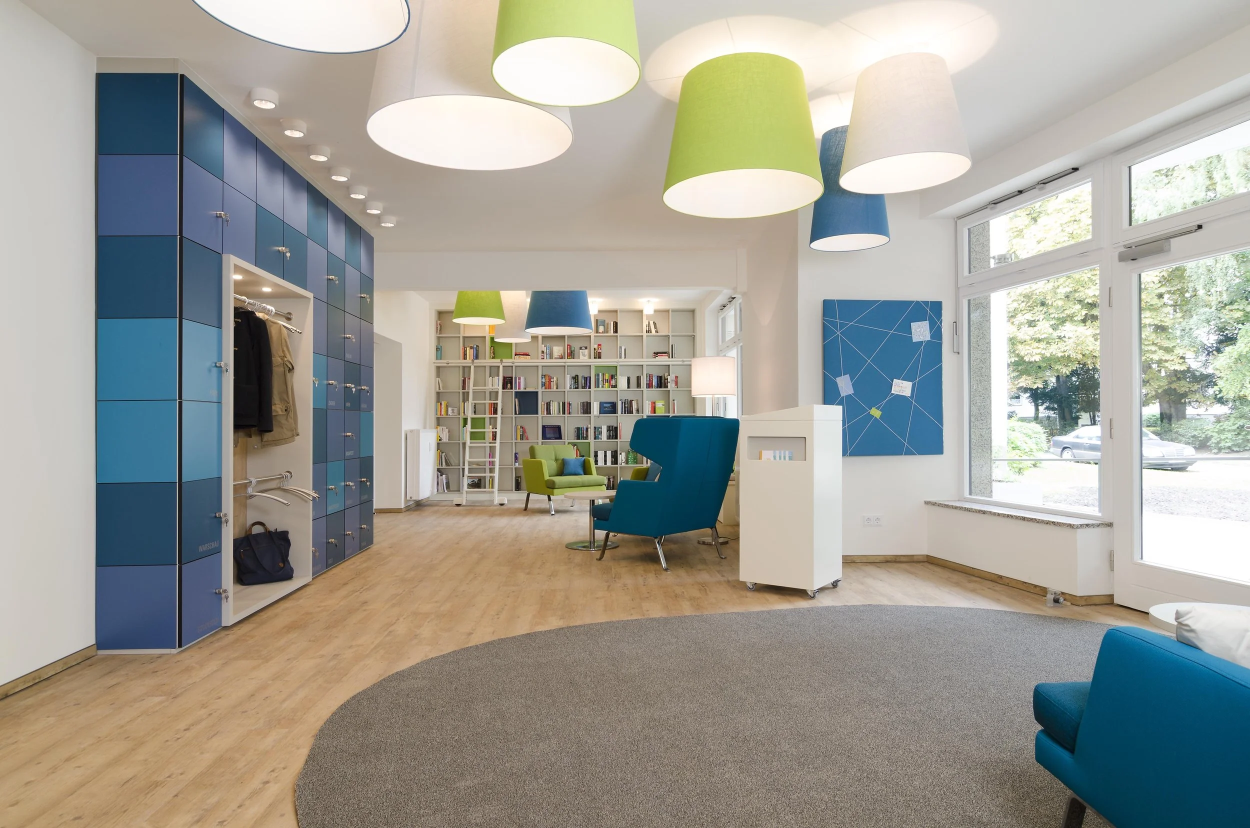 Moderner Aufenthaltsraum mit blauen, grünen und weißen Sitzmöbeln, großen bunten Deckenleuchten, Bücherregal im Hintergrund, Garderobenschränken auf der linken Seite, großem Fenster nach draußen, kleiner weißer Infostand, Wanddekoration, Bogen aus Te