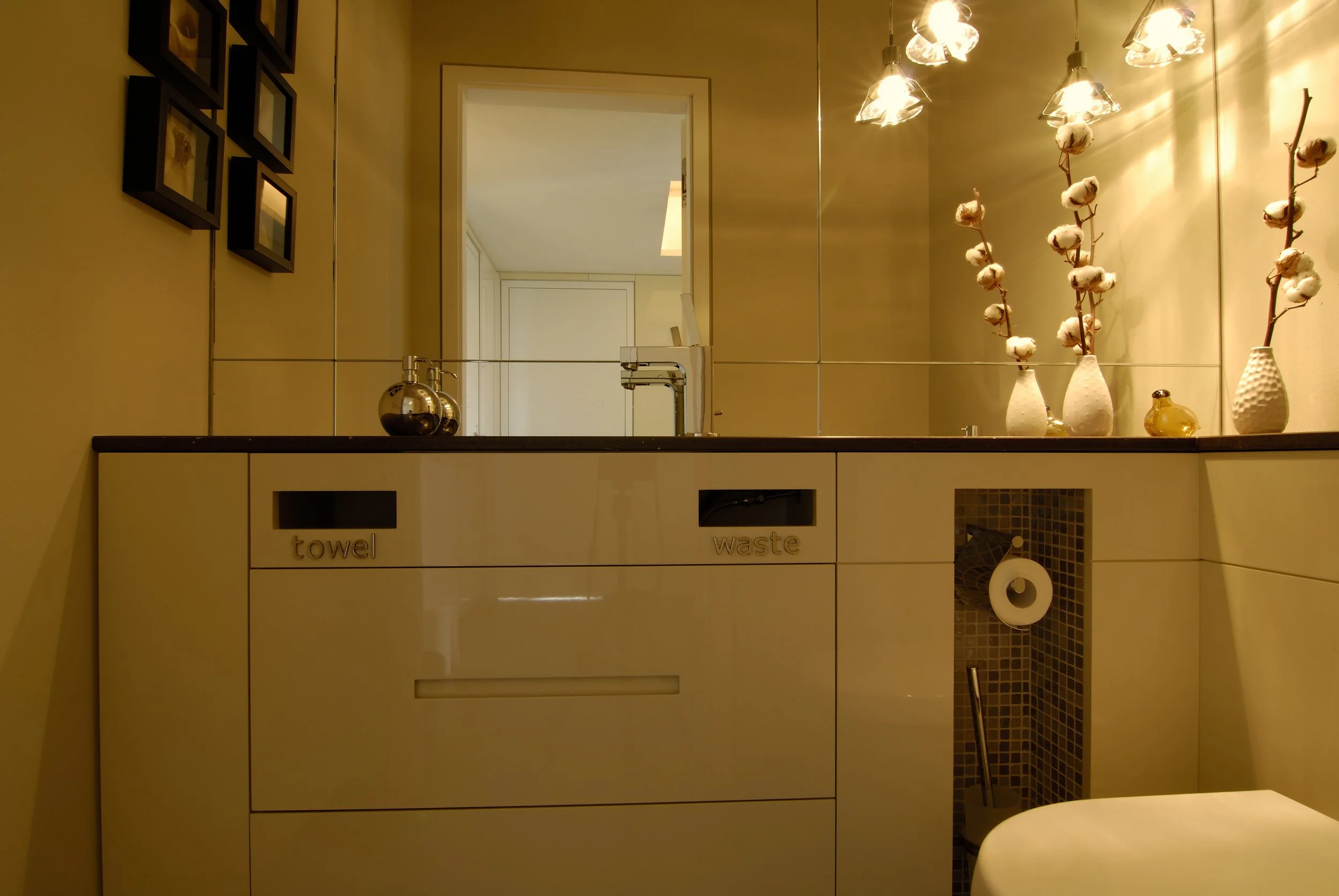 Helles Badezimmer mit Spiegel, Vasen mit Baumwollzweigen, Waschbecken, und einer Toilette, beleuchtet mit warmen Licht.