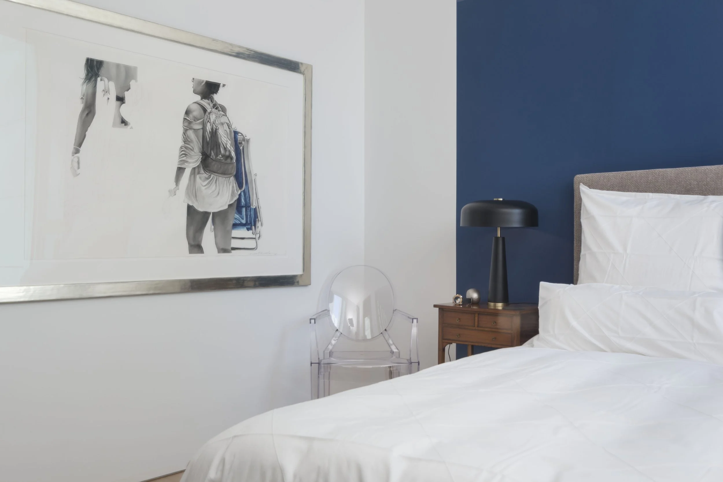 Ein modernes Schlafzimmer mit weißem Bett, dunkler blauer Wand, Nachttisch mit Schwarzlampe, Wandbild eines Menschen mit Rucksack, transparentem Stuhl und einem runden Spiegel.