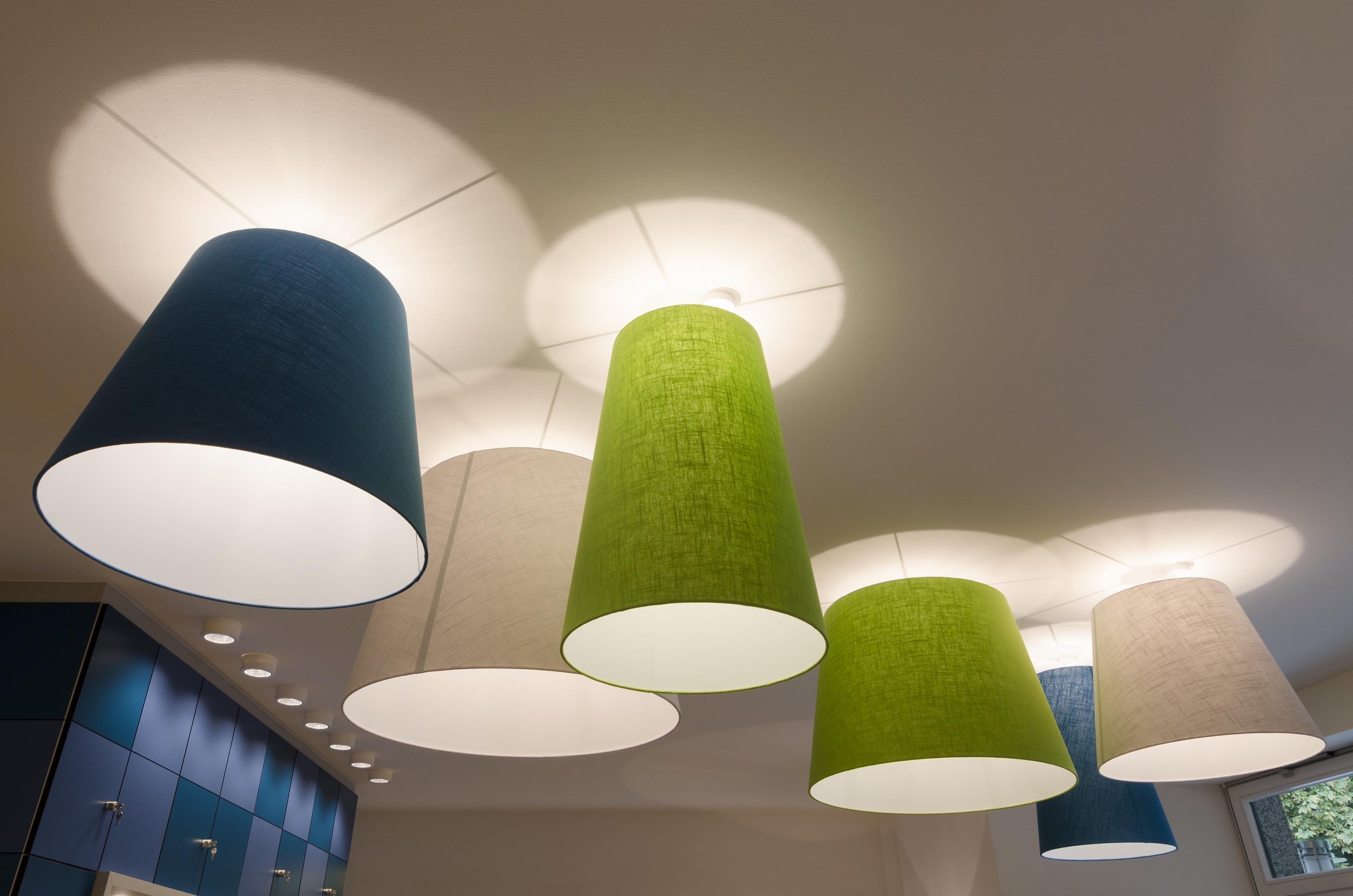 Mehrere bunte Lampenschirme in Blau, Grün, Beige und Weiß hängen von der Decke eines Raums, beleuchten den Raum und werfen Schatten an die Decke.