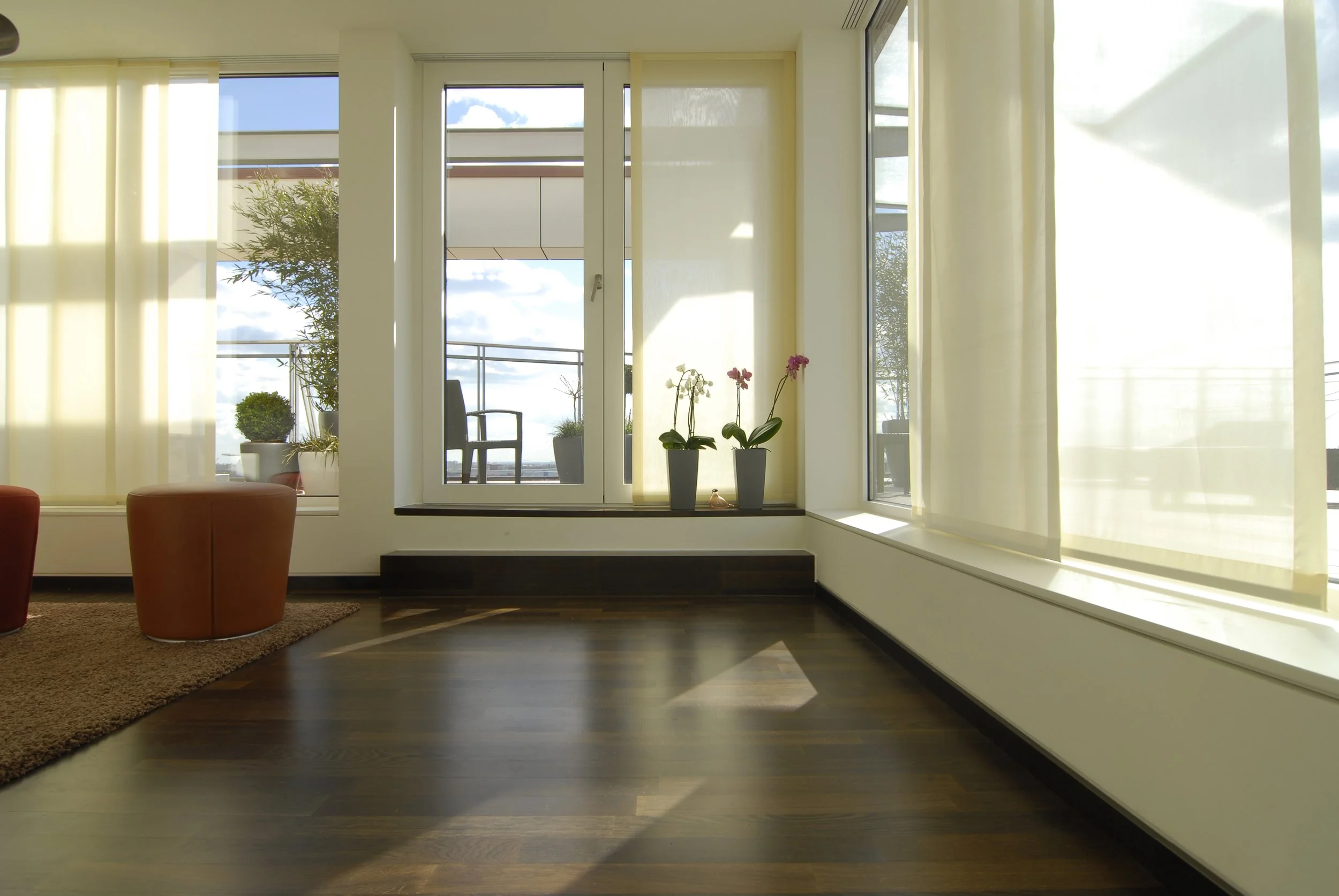 Heller, moderner Wohnzimmerbereich mit dunklem Holzparkett, großen Fenstern mit Vorhängen, einigen Pflanzen auf der Fensterbank, einer Gartenstühle auf dem Balkon im Hintergrund.