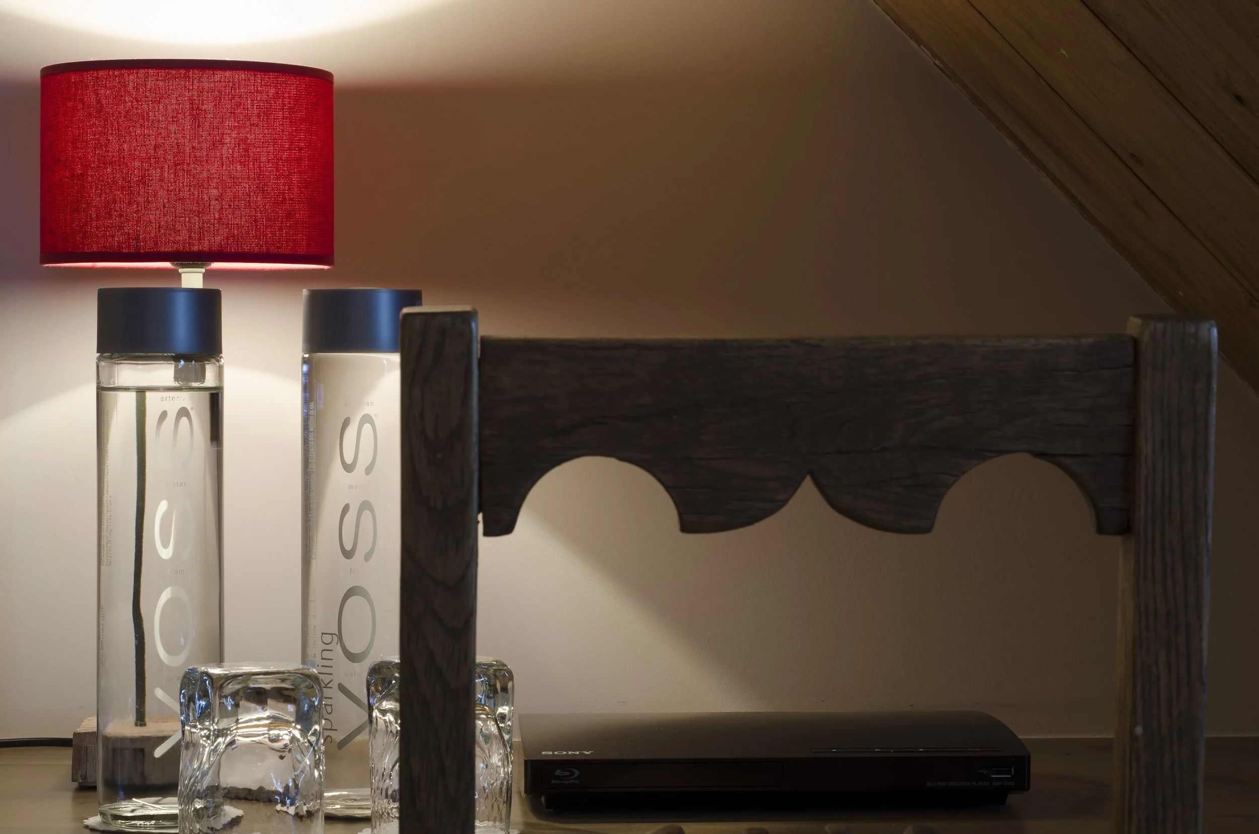 Zwei Wasserflaschen mit schwarzen Deckeln und die Beschriftung 'Water' sowie 'S' auf einer Holzoberfläche, rote Tischlampe, zwei leere Gläser umgekippt, ein schwarzer Blu-ray Player, im Hintergrund eine Wand und eine Dachschräge.