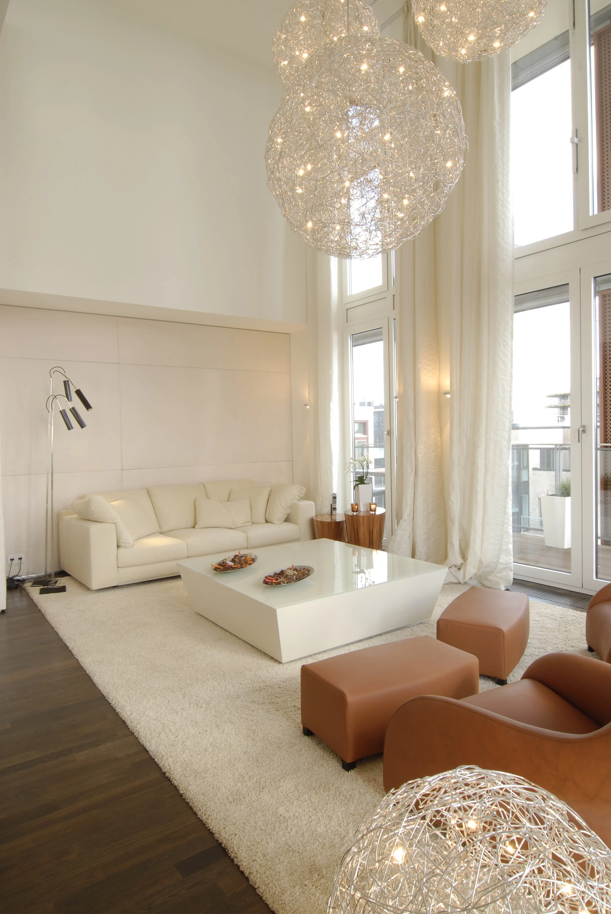 Modernes Wohnzimmer mit hohen Fenstern, weißen Vorhängen, einer weißen Couch, braunen Sessel, einem weißen Couchtisch, einer Stehlampe und einer Deckenleuchte aus Korb. Der Raum ist hell und stilvoll eingerichtet.
