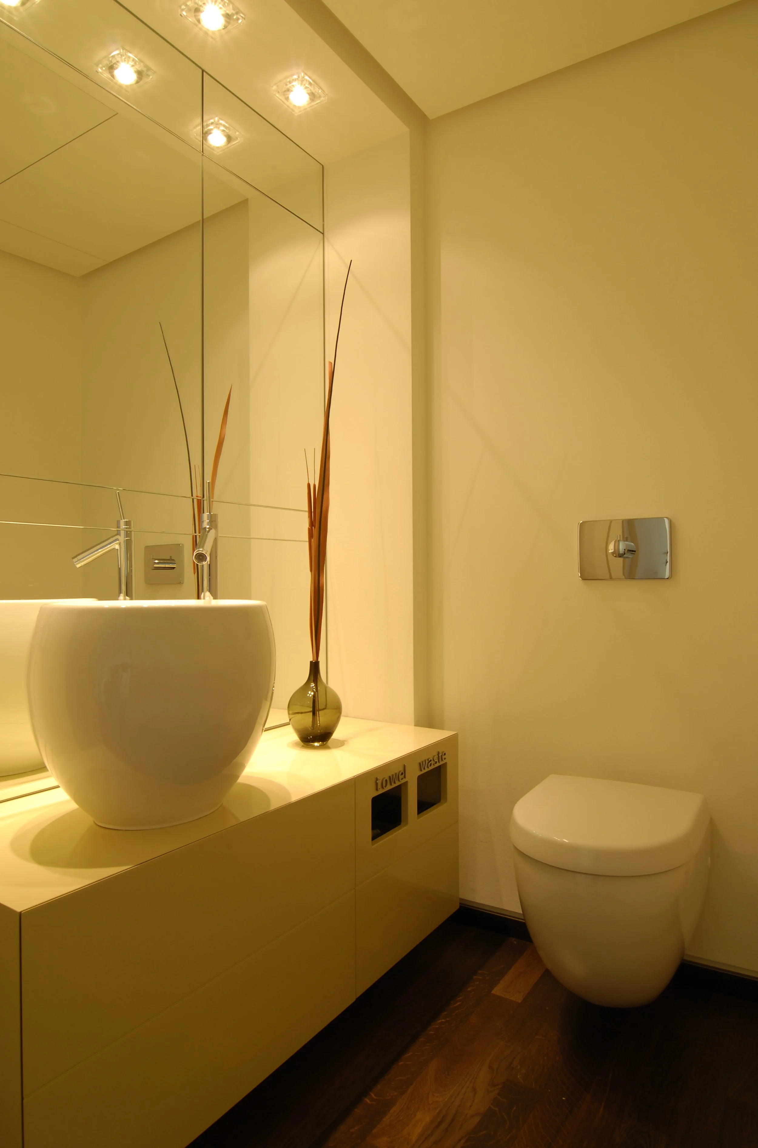 Moderne Badezimmer mit einer weißen runden Waschschüssel, einem großen Spiegel und einer Vase mit Dekoknospen, eine weiße Toilette, dunkler Holzboden.