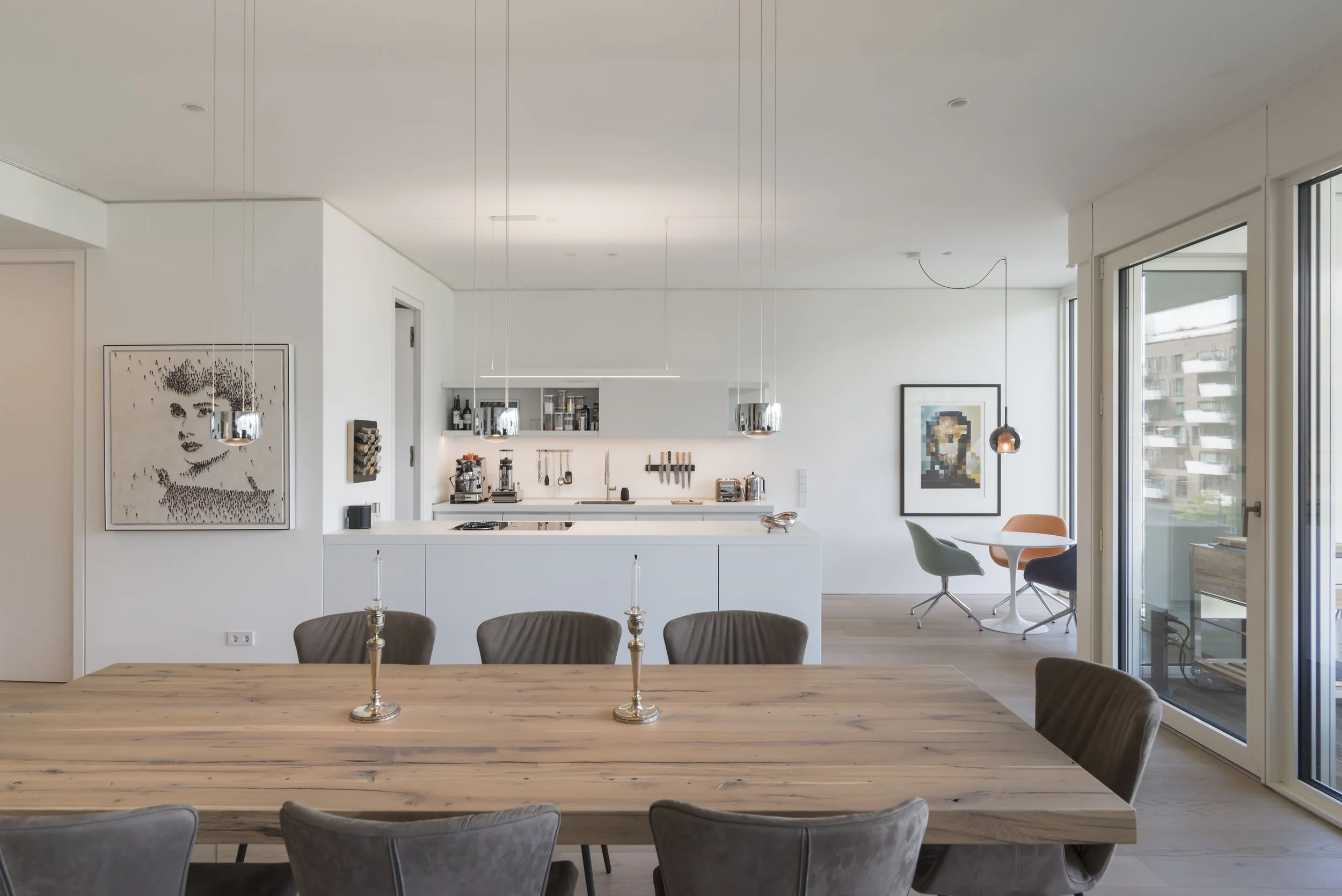 Offene, moderne Küche und Essbereich mit großem Holztisch und Stühlen, große Fensterfronten, Kunstwerke an den Wänden, minimalistische Beleuchtung, Balkon sichtbar im Hintergrund.