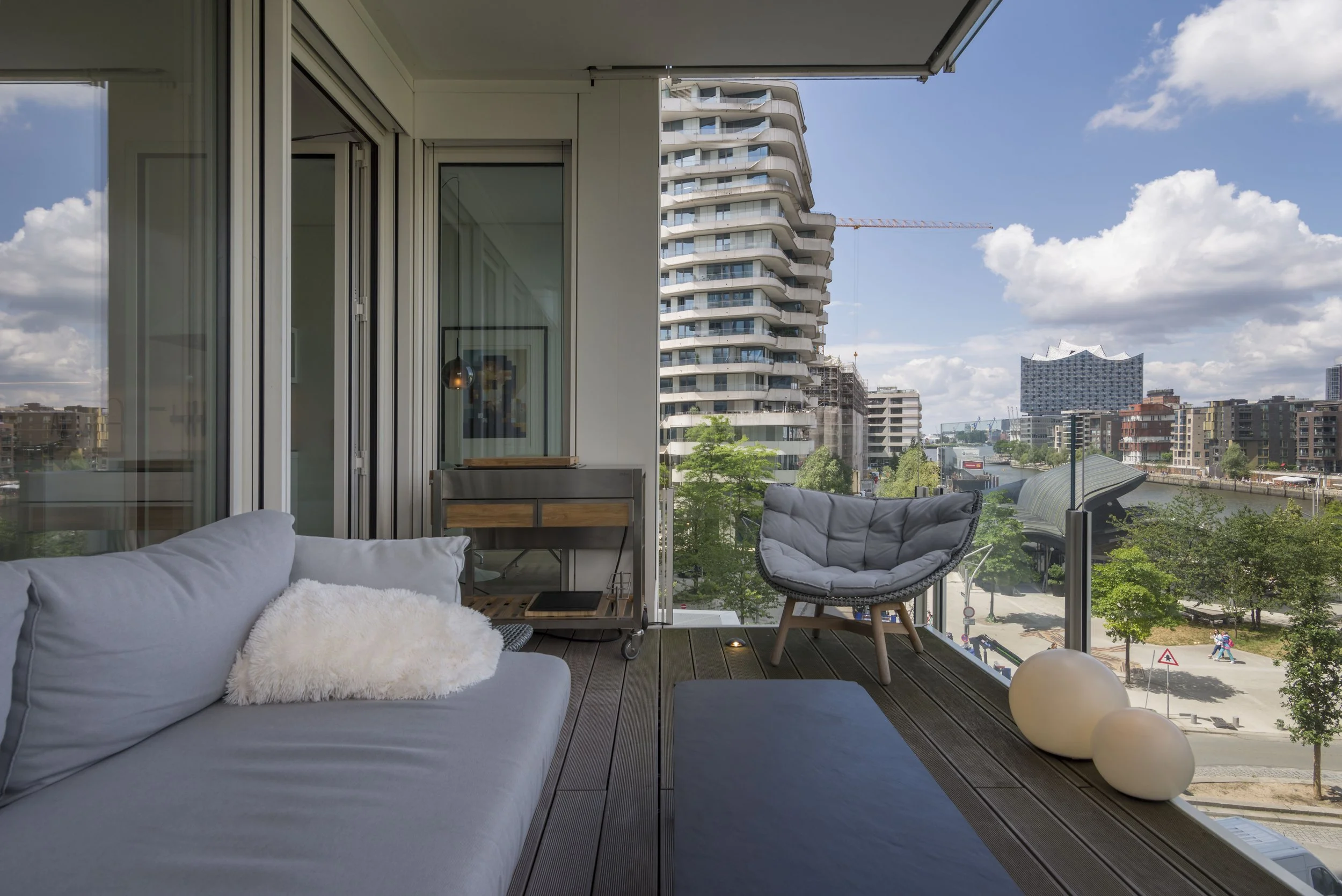 Balkon mit grauem Sofa, einem modernen Sessel, weißen Dekokugeln und Blick auf Stadt mit hohen Gebäuden, Wasser und Kinderspielplatz bei schönem Wetter.