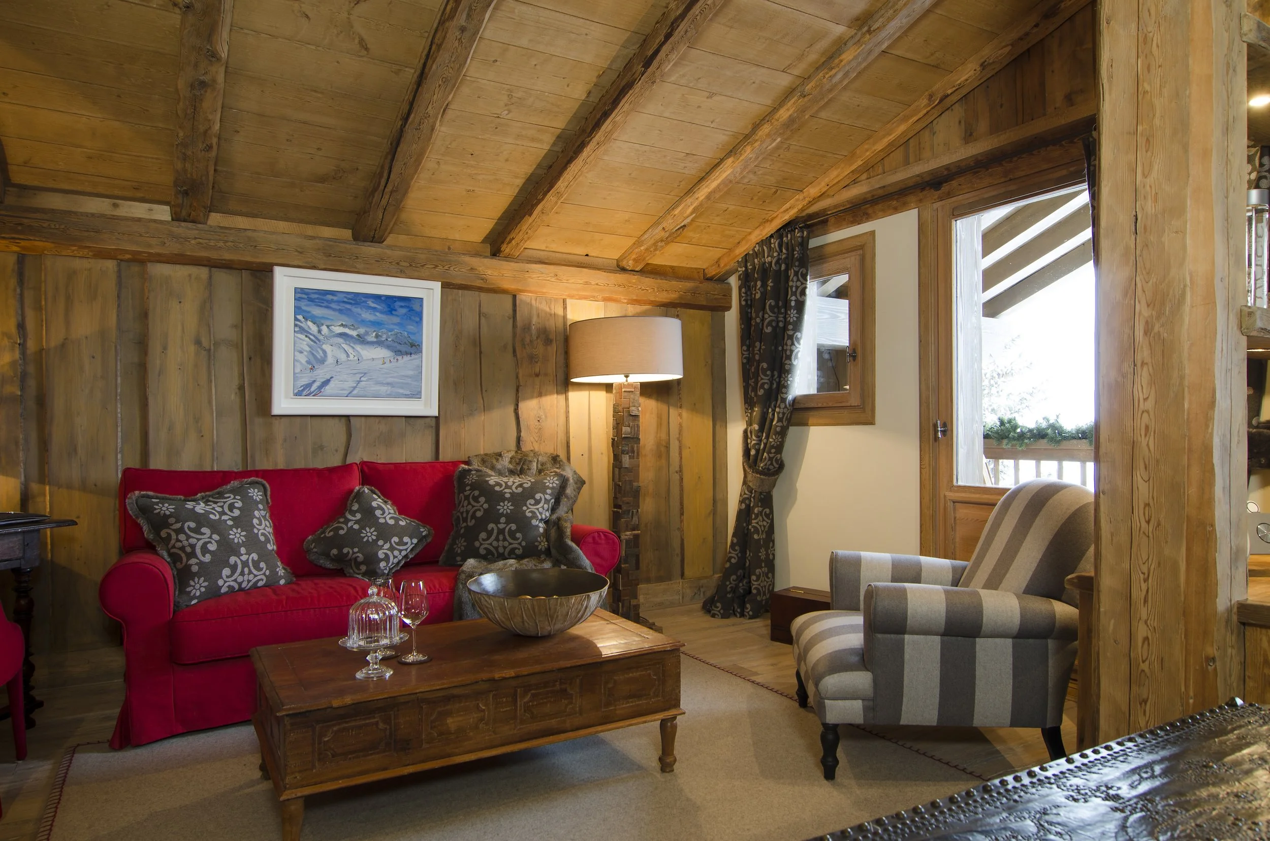 Gemütliches Wohnzimmer mit roten und gepunkteten Kissen, Holzmöbeln, einem Bild eines verschneiten Berglands, einer Stehlampe, einem Fenster und einer Tür, die auf eine Terrasse mit Holzgeländer führt.