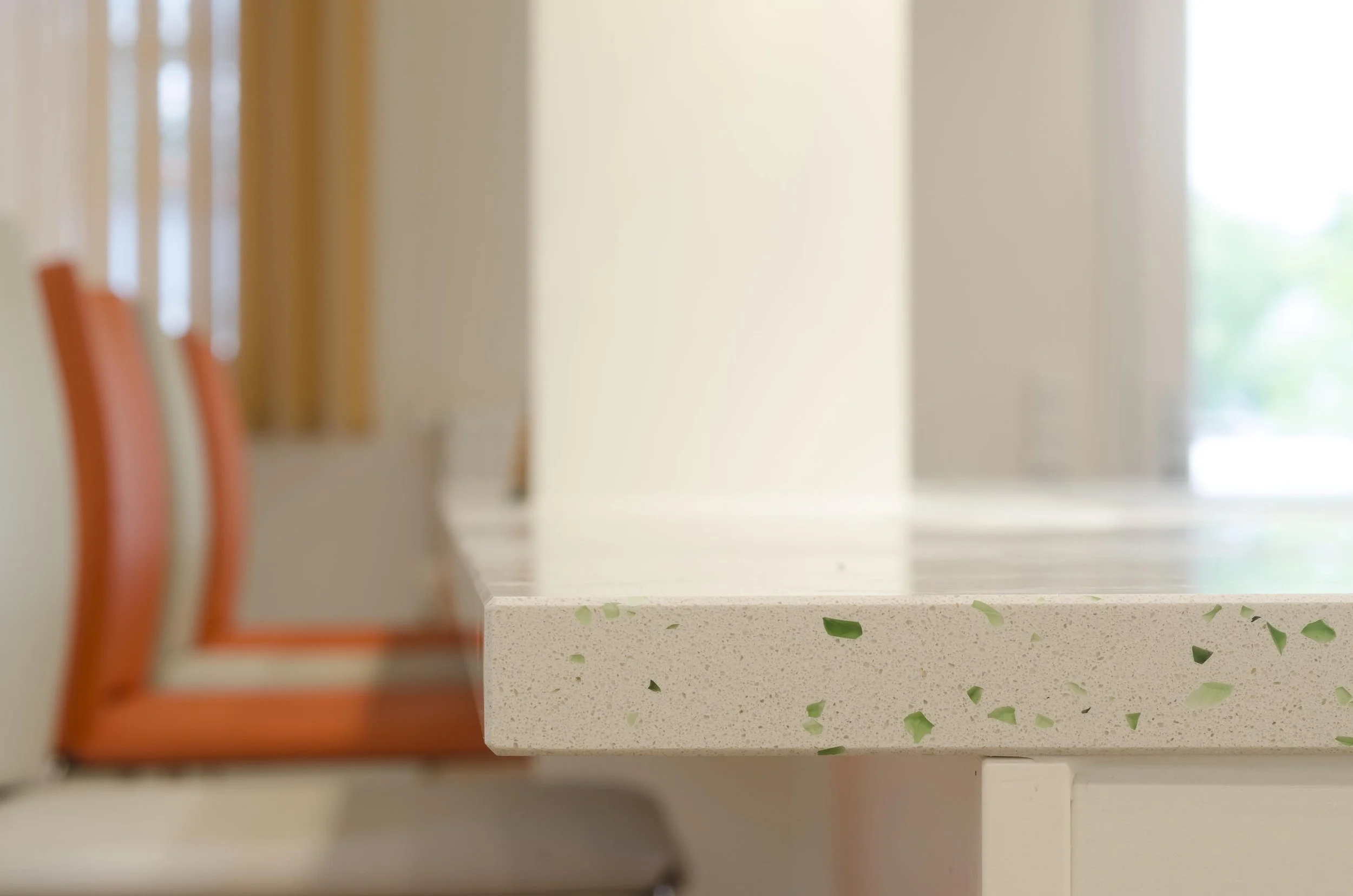 Nahaufnahme einer weißen Küchenarbeitsplatte mit grünen Steinchen, im Hintergrund eine helle Küche mit großen Fenstern.