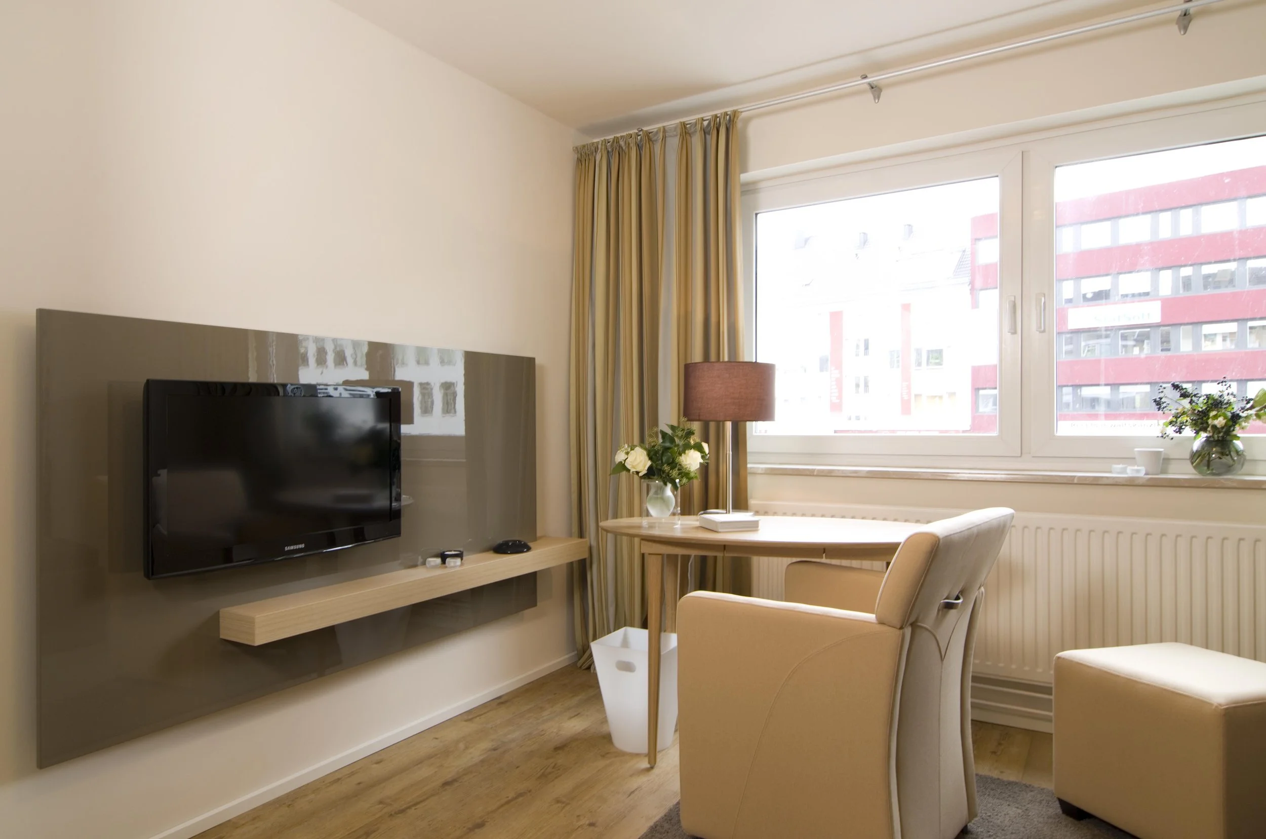 Ein gemütliches Hotelzimmer mit einem Flachbildfernseher, einem kleinen Schreibtisch mit Lampe und Blumen, einem bequemen Sessel, einem Fenster mit blick auf ein urbanes Gebiet, und einer kleinen Vase mit Blumen auf der Fensterbank.