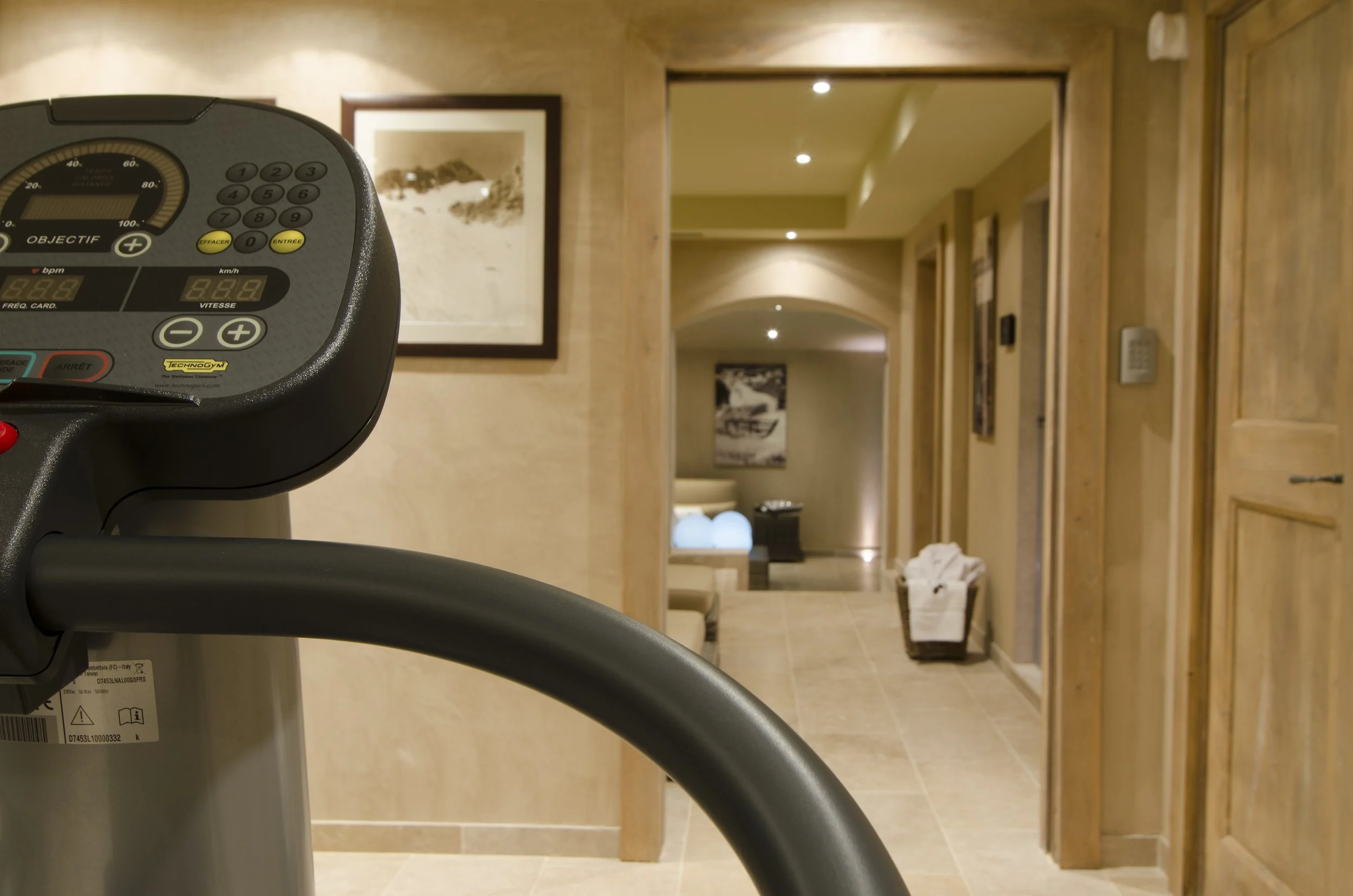 Ein Heimtrainer im Vordergrund, Blick in einen hell erleuchteten Flur eines Hauses mit Bilder an den Wänden und einem Korb mit weißen Handtüchern.