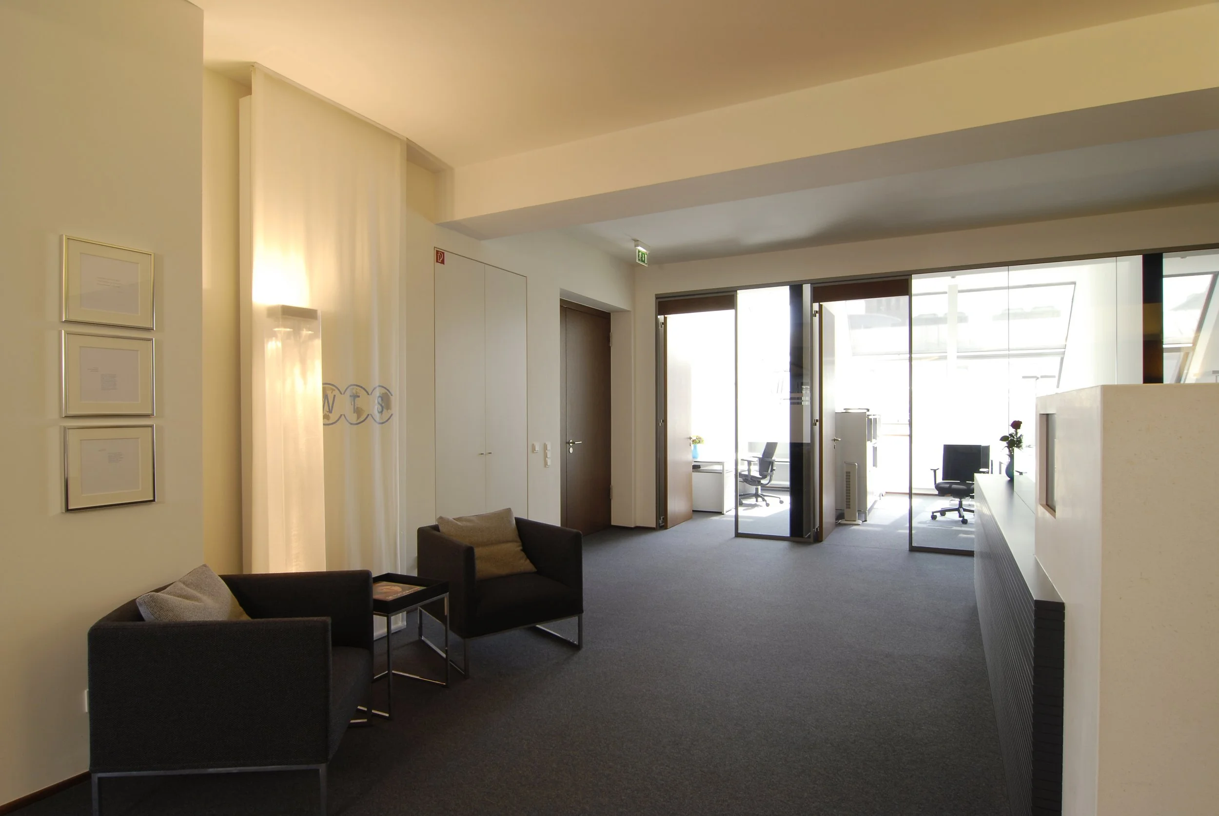 Wartezimmer mit zwei schwarzen Sesseln, einem kleinen Tisch, großen Fenstern und einem Arbeitsbereich mit Stühlen im Hintergrund.