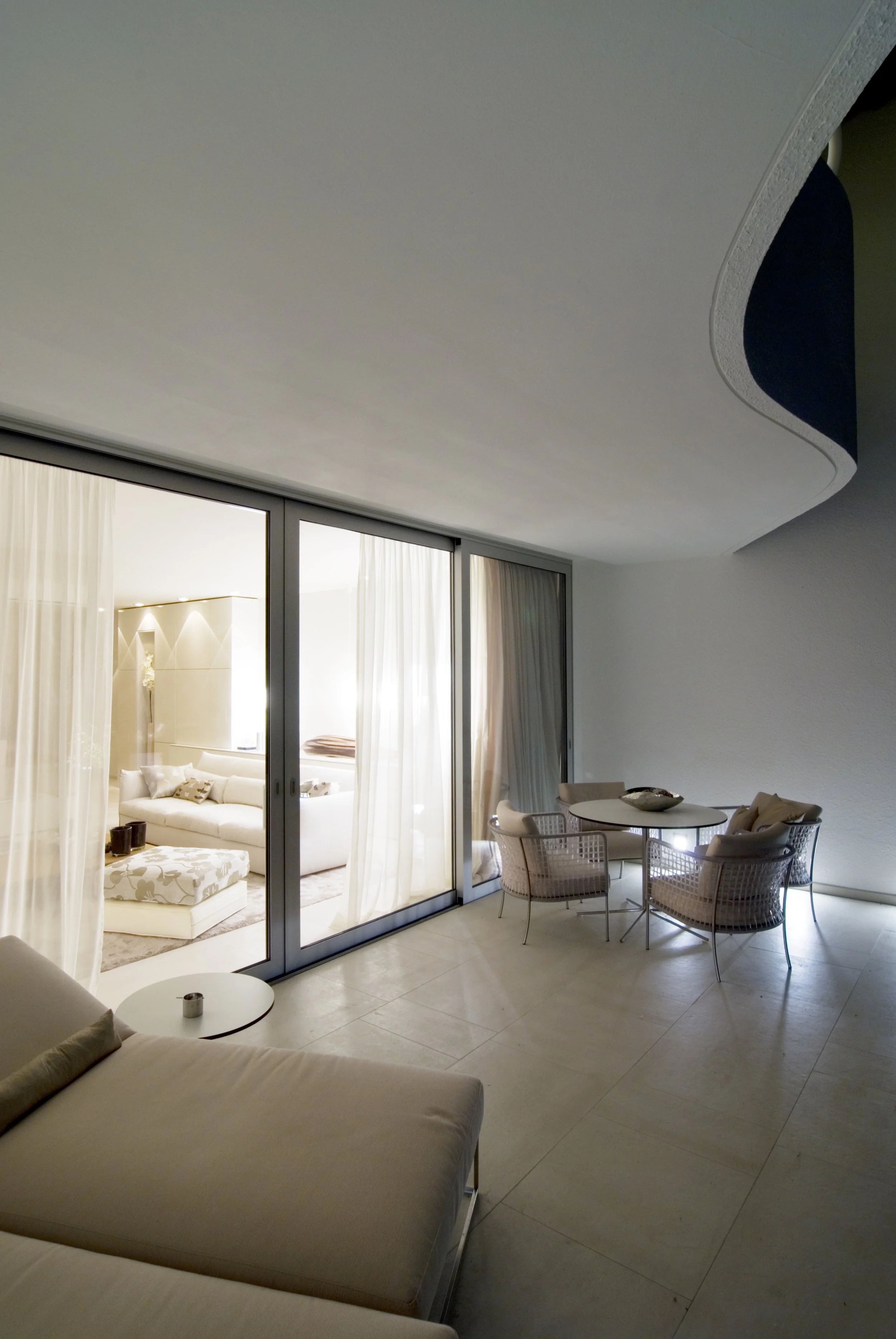 Moderner Wohnraum mit offener Terrasse, Sitzbereich mit Sofas, Tisch und Stühlen, helle Wände und große Glasfenster mit Vorhängen.