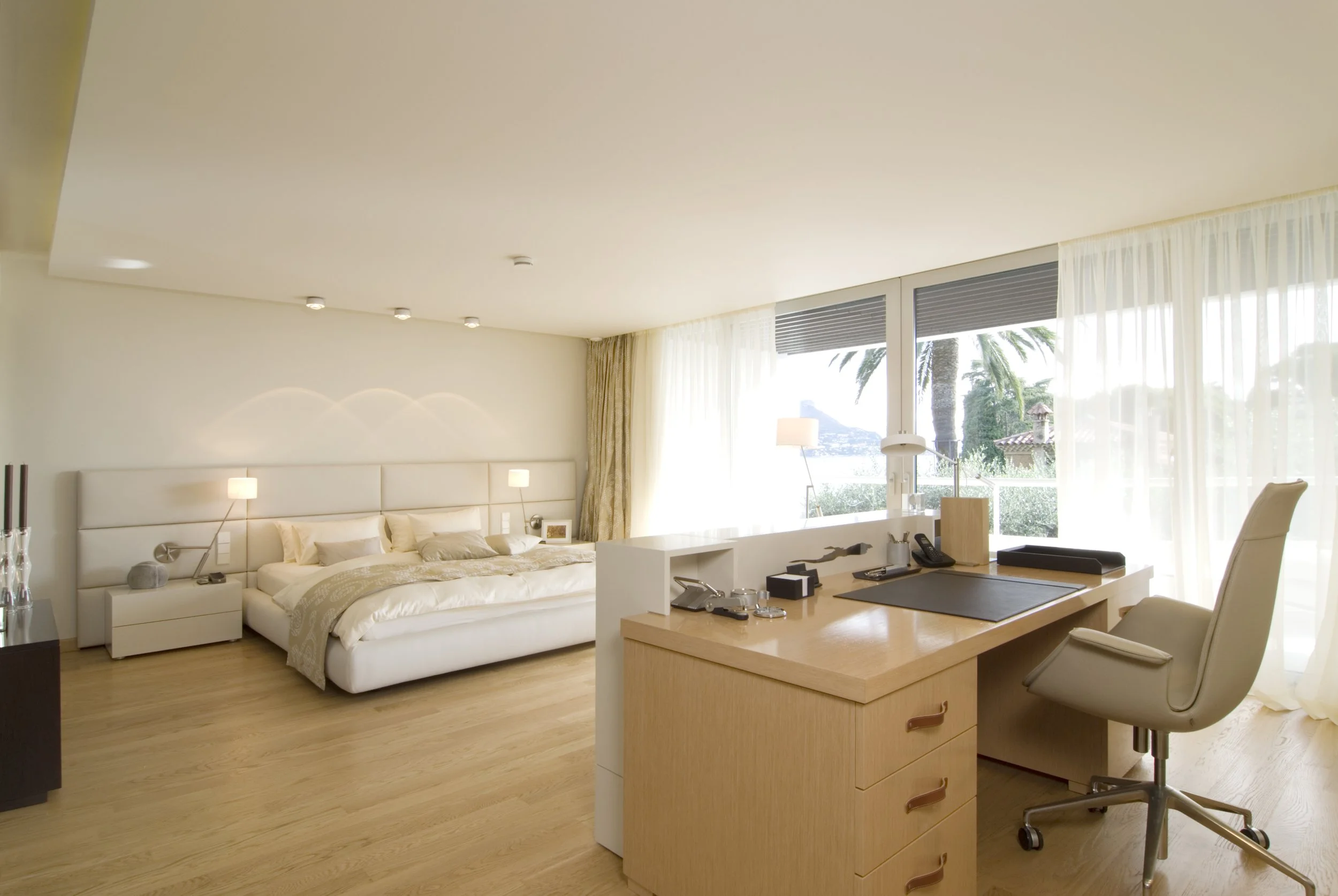 Ein modernes Hotelzimmer mit einem großen Bett, Nachttischen, einem Schreibtisch mit Stuhl und einer großen Fensterfront mit Blick auf Palmen im Außenbereich.