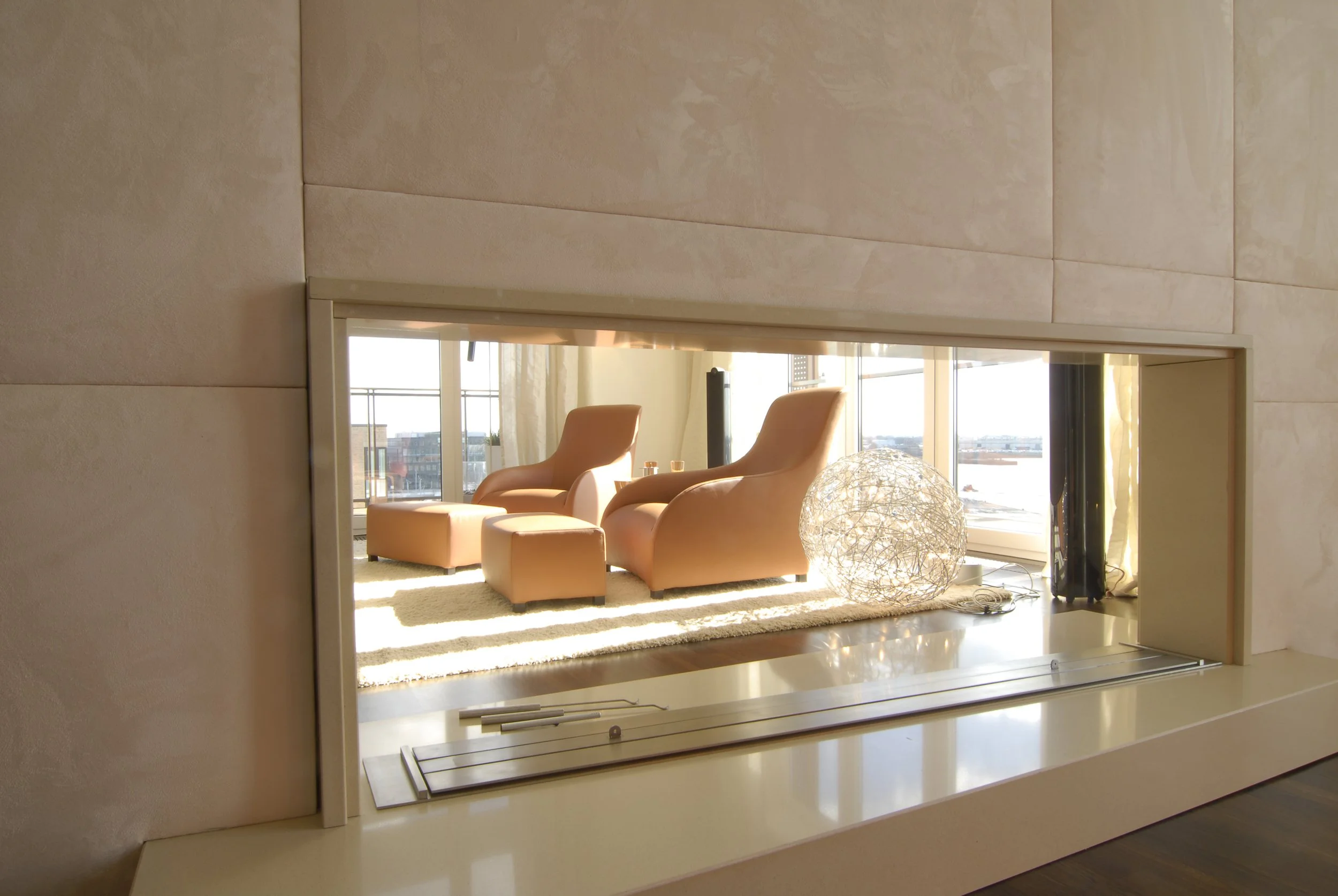 Ein Wohnzimmer mit zwei hellbraunen Sessel, einem Fußhocker, einem großen sphericalen Lampenschirm und einem großen Fenster mit Blick auf die Skyline.