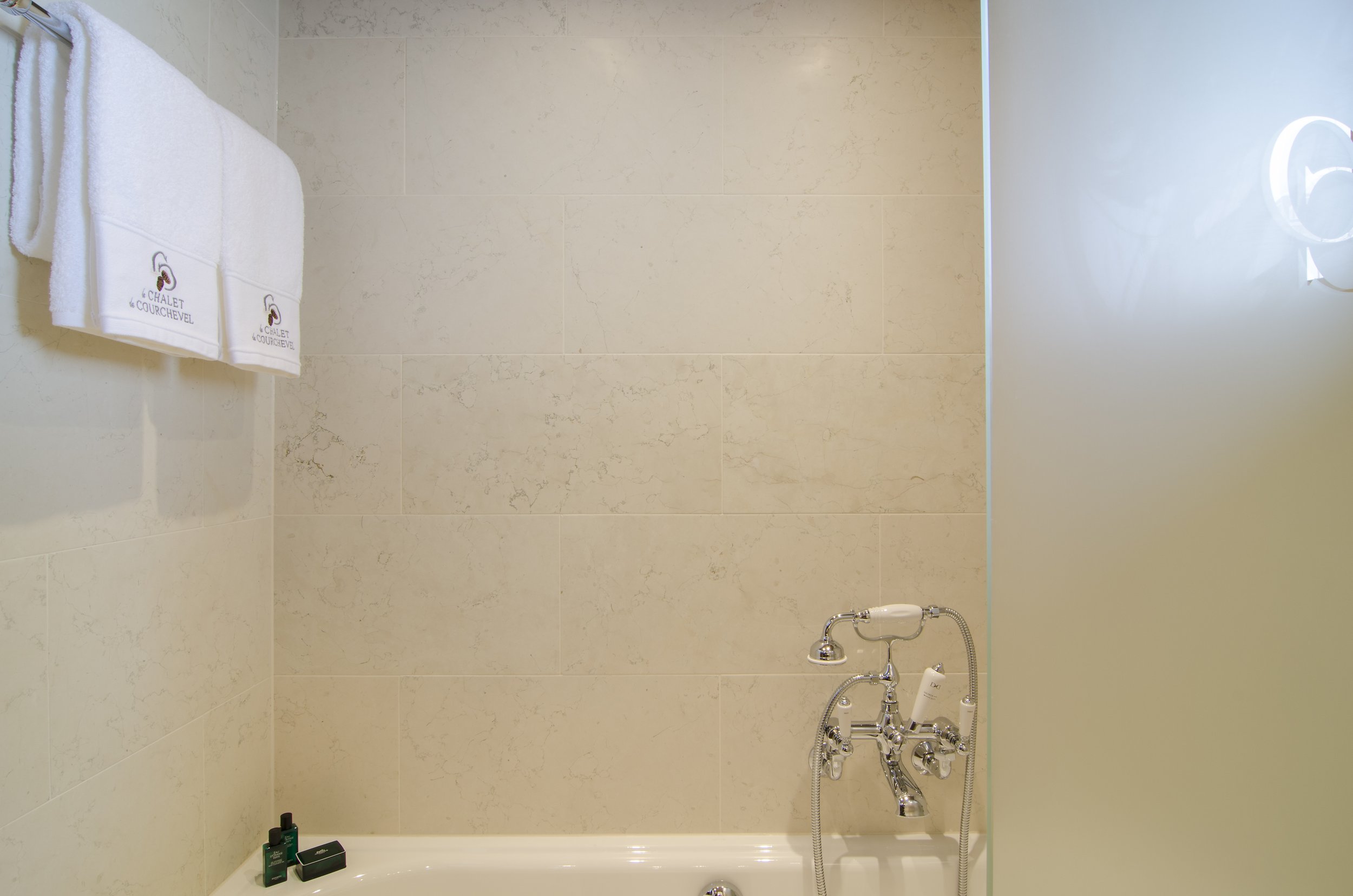 Badezimmer mit weißen Handtüchern, Shampoo und Seife auf der Badewanne, Wasserhahn und Dusche, cremefarbene Fliesen an der Wand, Teil eines gläsernen Badezimmerscheiben.