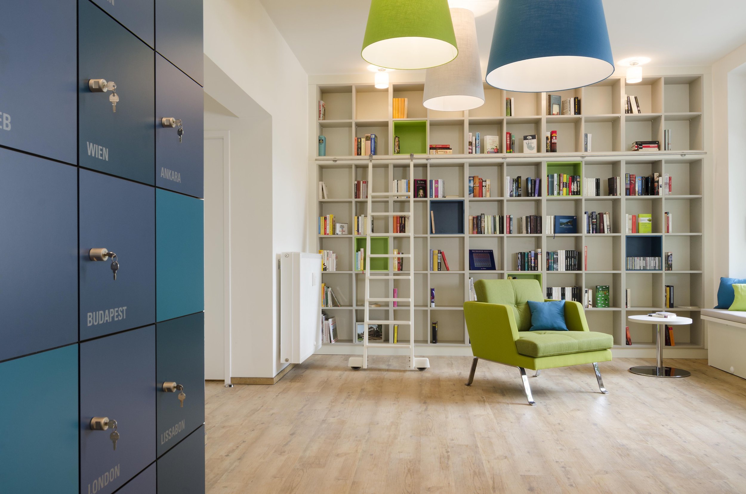 Moderner Raum mit blauen Spindern auf der linken Seite, weißen Bücherregalen an der Wand, grüner Sessel mit Kissen und kleinen Tischen. Die Decke hat bunte Lampen in grün, beige, grau und blau. Der Raum hat Holzfußboden und viel Tageslicht.