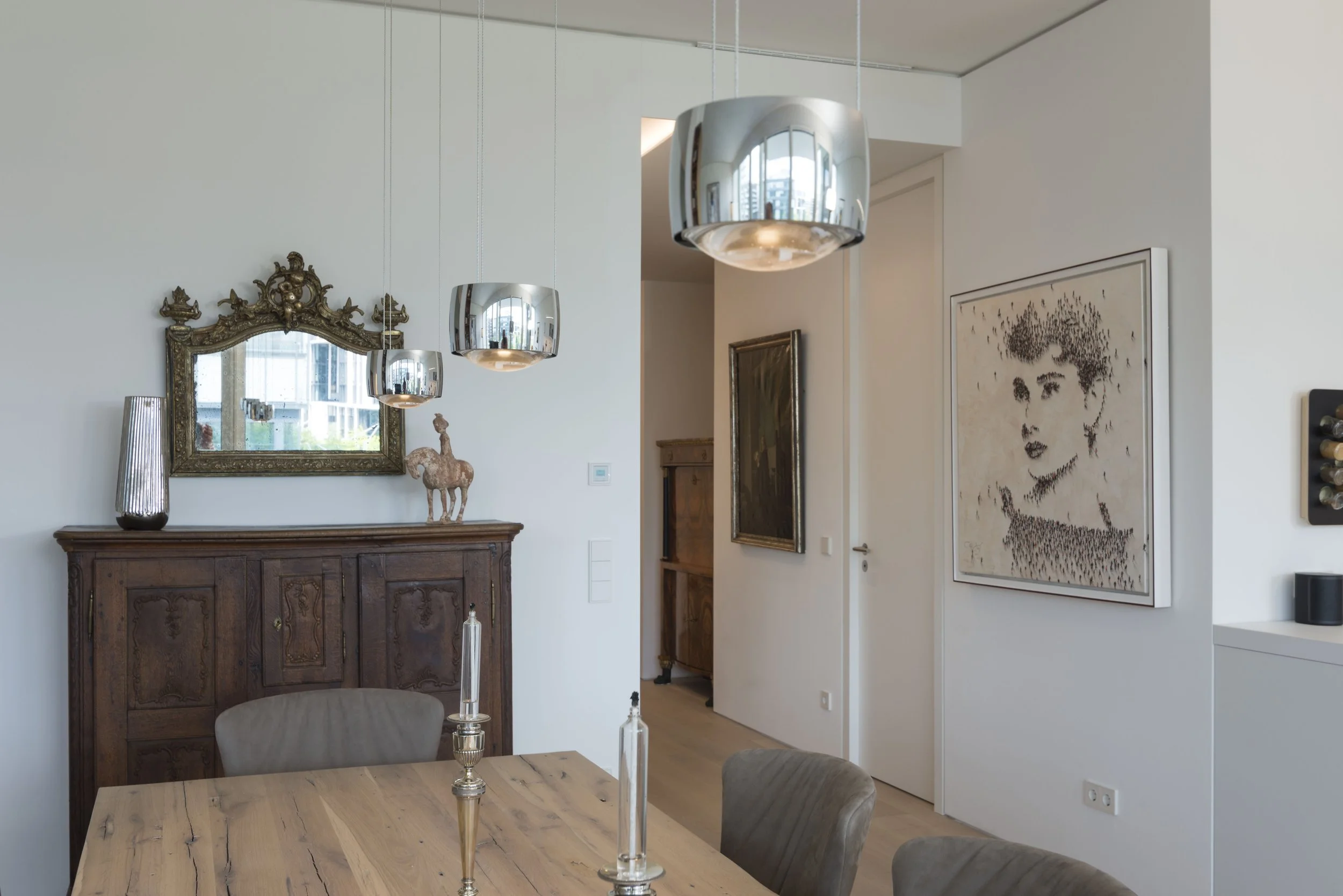 Moderner Essbereich mit Vintage- und modernen Dekorationen, darunter große silberne Hängeleuchten, ein antiker Holschrank, ein Wandspiegel, eine Plüsch-Giraffe, Gemälde und eine Wand mit Rotweinregal.