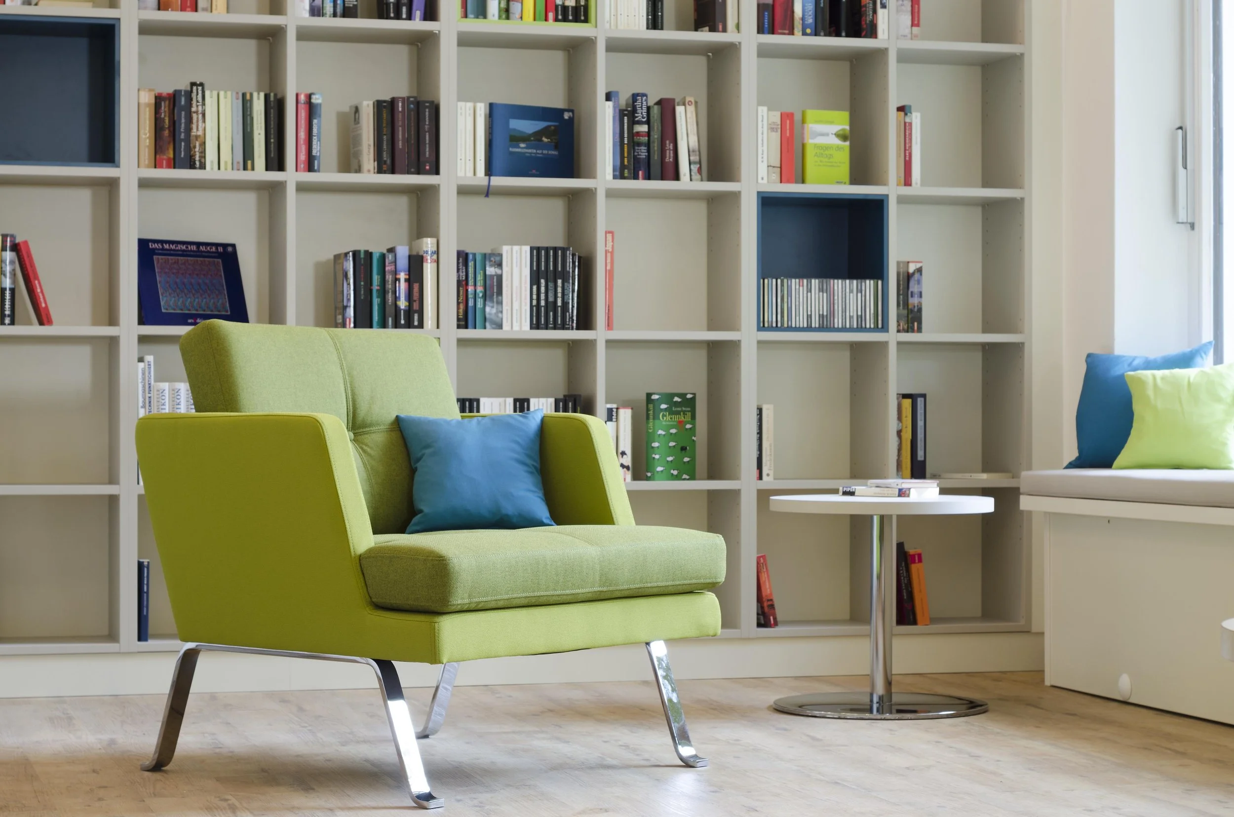 Moderner Leseraum mit grüner Sessel, Regal mit Büchern im Hintergrund und Fenster auf der rechten Seite.