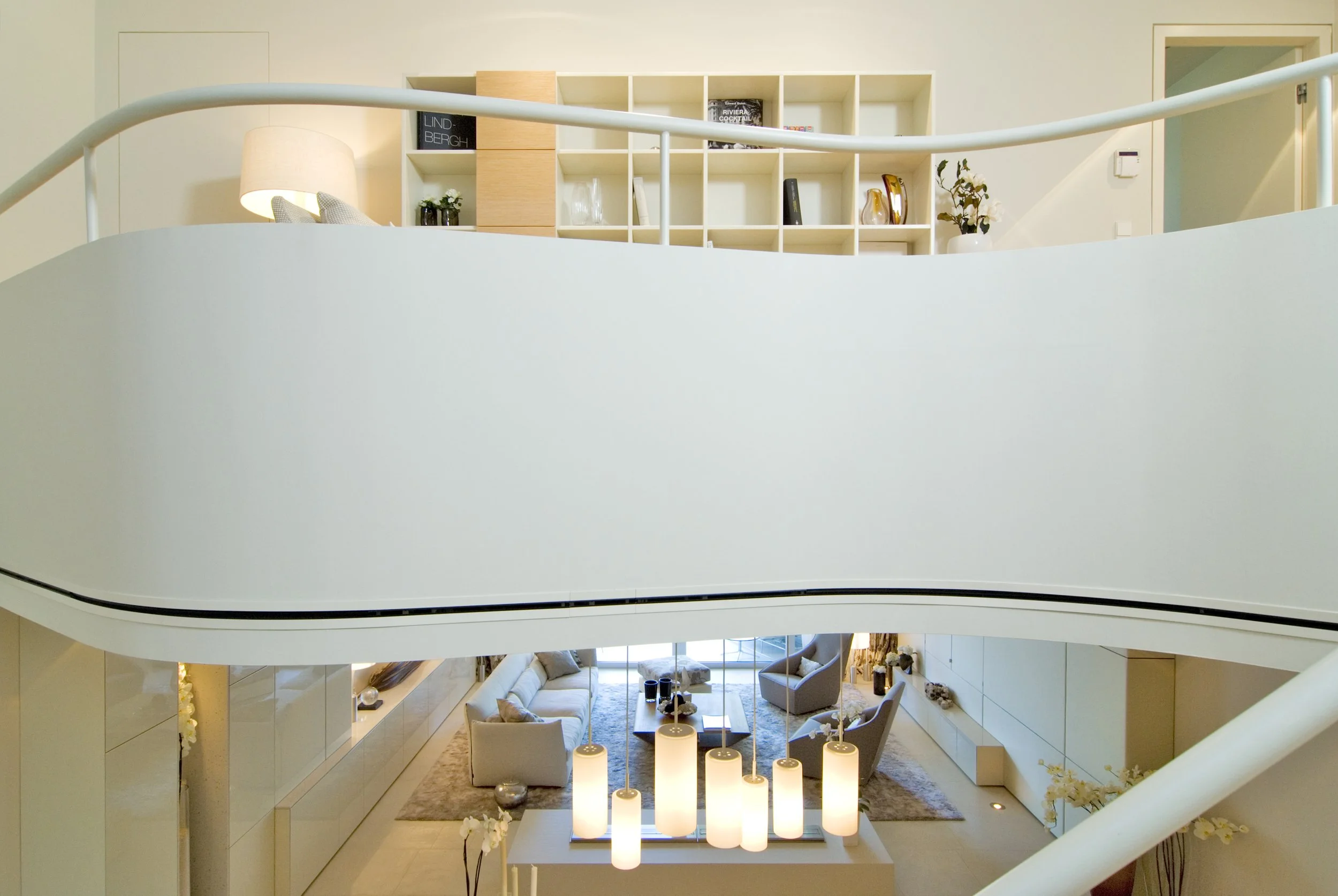 Ein Blick auf ein modernes, offen gestaltetes Wohnzimmer mit Sitzecke, weißen Möbeln, einem Teppich, Wandregalen und stylischen Lampen, aufgenommen von einer oberen Etage.