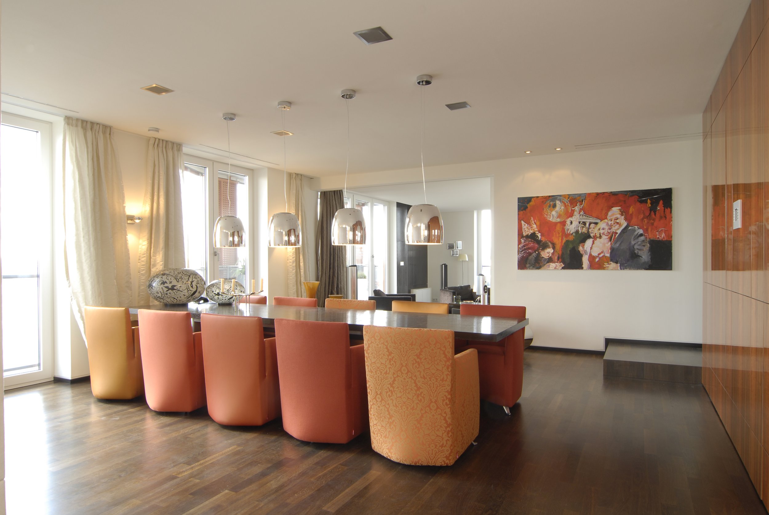 Ein modernes Esszimmer mit einem großen rechteckigen Tisch, umgeben von mehreren bunten Stühlen in Orangetönen. Es gibt vier Kronleuchter über dem Tisch, große Fenster mit weißen Vorhängen und eine bunte Wandmalerei an der Wand. Hinten im Raum ist ei