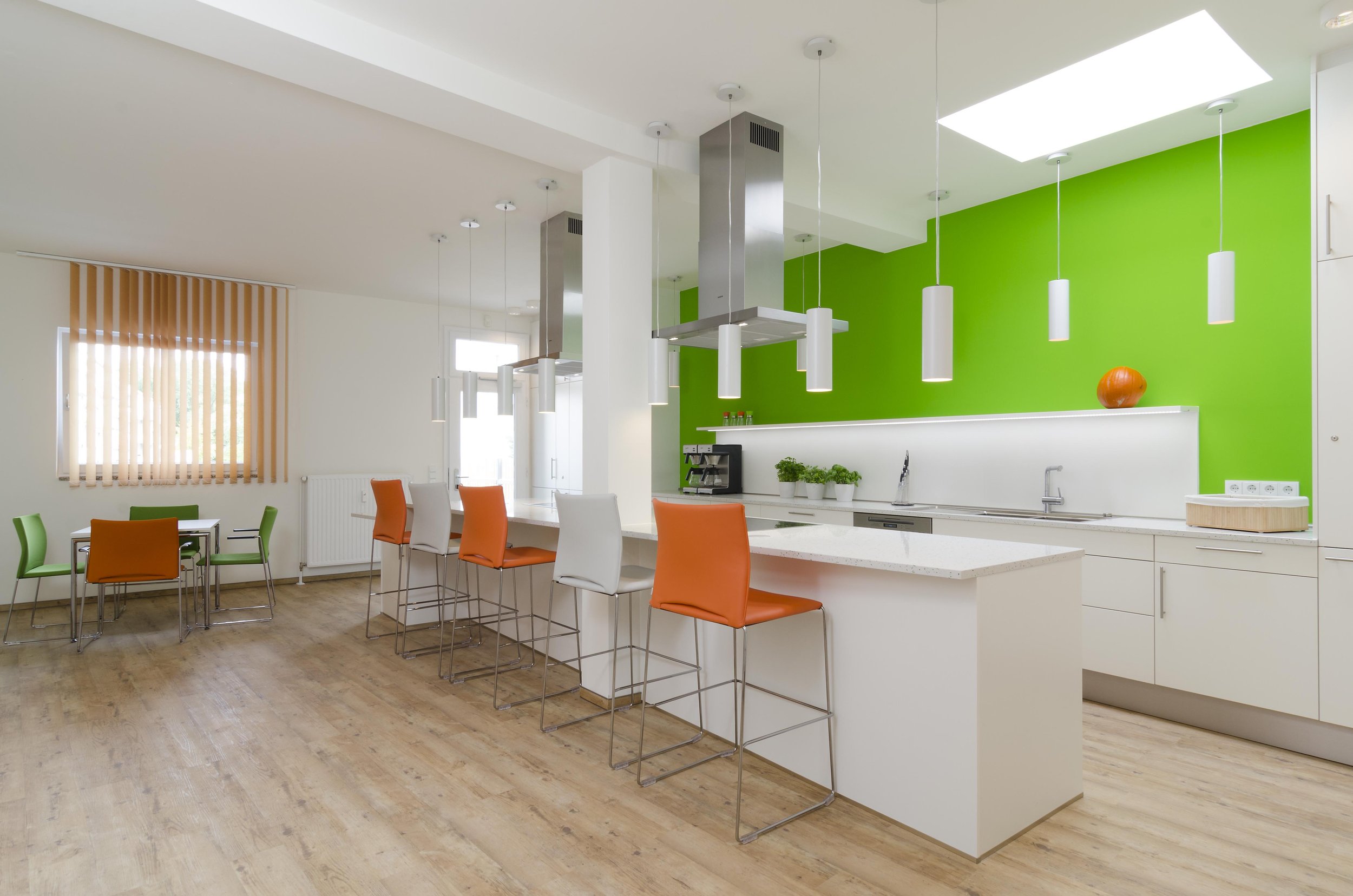 Moderne, helle Küche mit einer weißen Kücheninsel, bunten Stühlen und einer grünen Wand im Hintergrund.