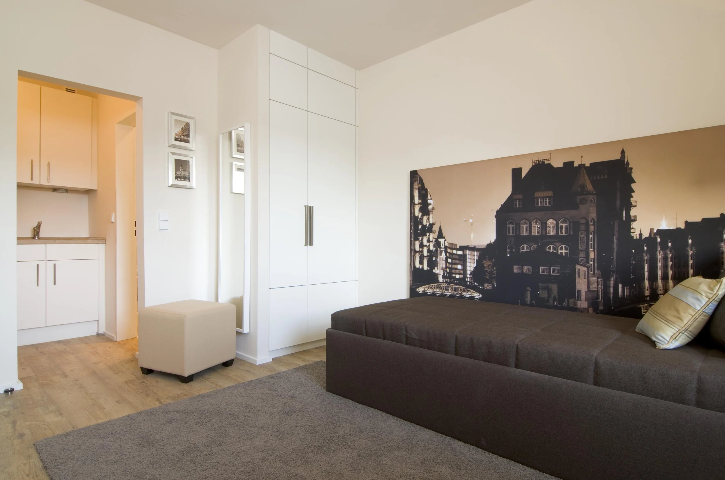 Ein modernes Schlafzimmer mit dunklem Bett, einer großflächigen Wandfotografie eines Stadtbildes bei Sonnenuntergang, einem weißen Kleiderschrank, einem kleinen weißen Hocker und einem beige-grauen Teppich.