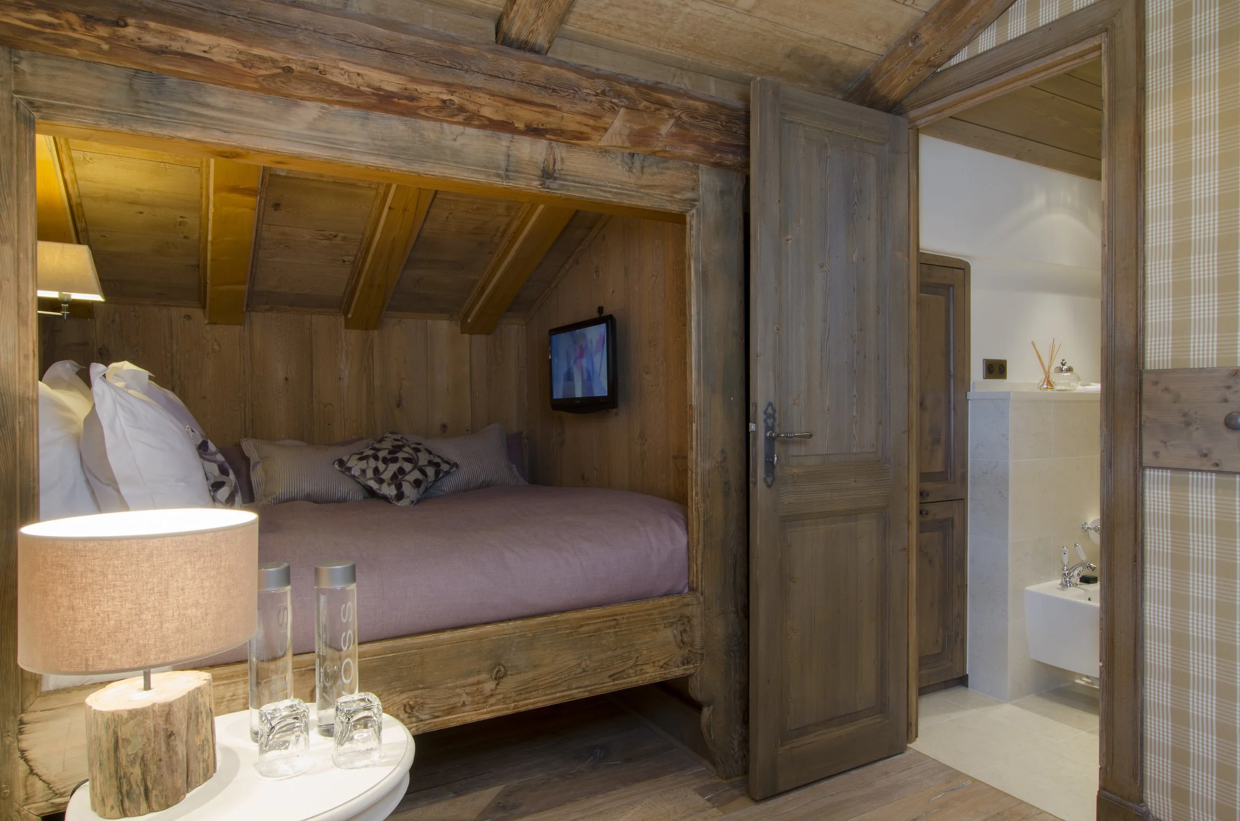 Ein Schlafzimmer in einer Holzhütte mit einem Bett mit rosa Bettwäsche, einer kleinen Lampe, einem Fernseher und Zugang zu einem Badezimmer.
