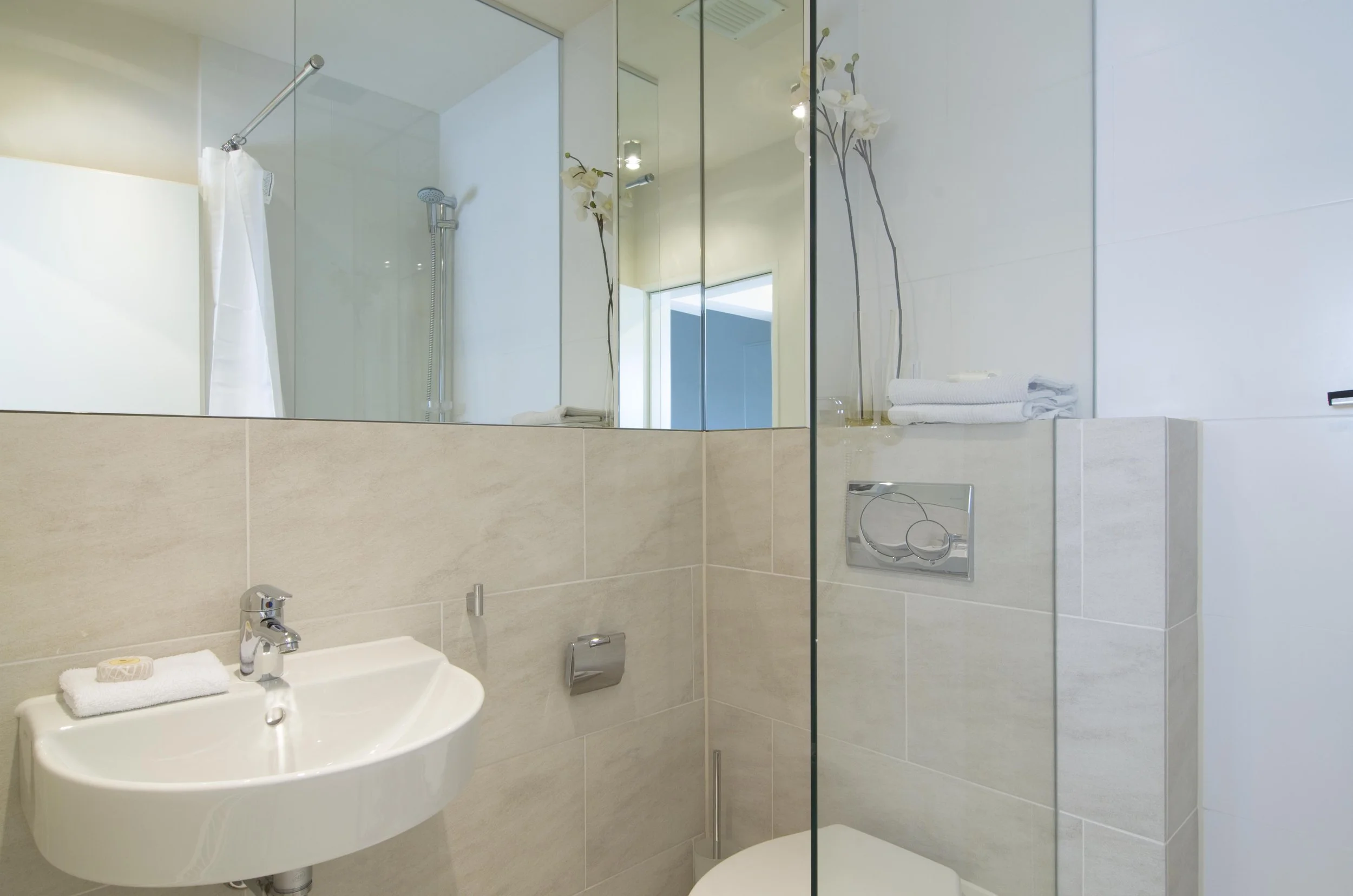 Modernes Badezimmer mit Dusche, Waschbecken, weißen Handtüchern und Orchideen im Hintergrund.