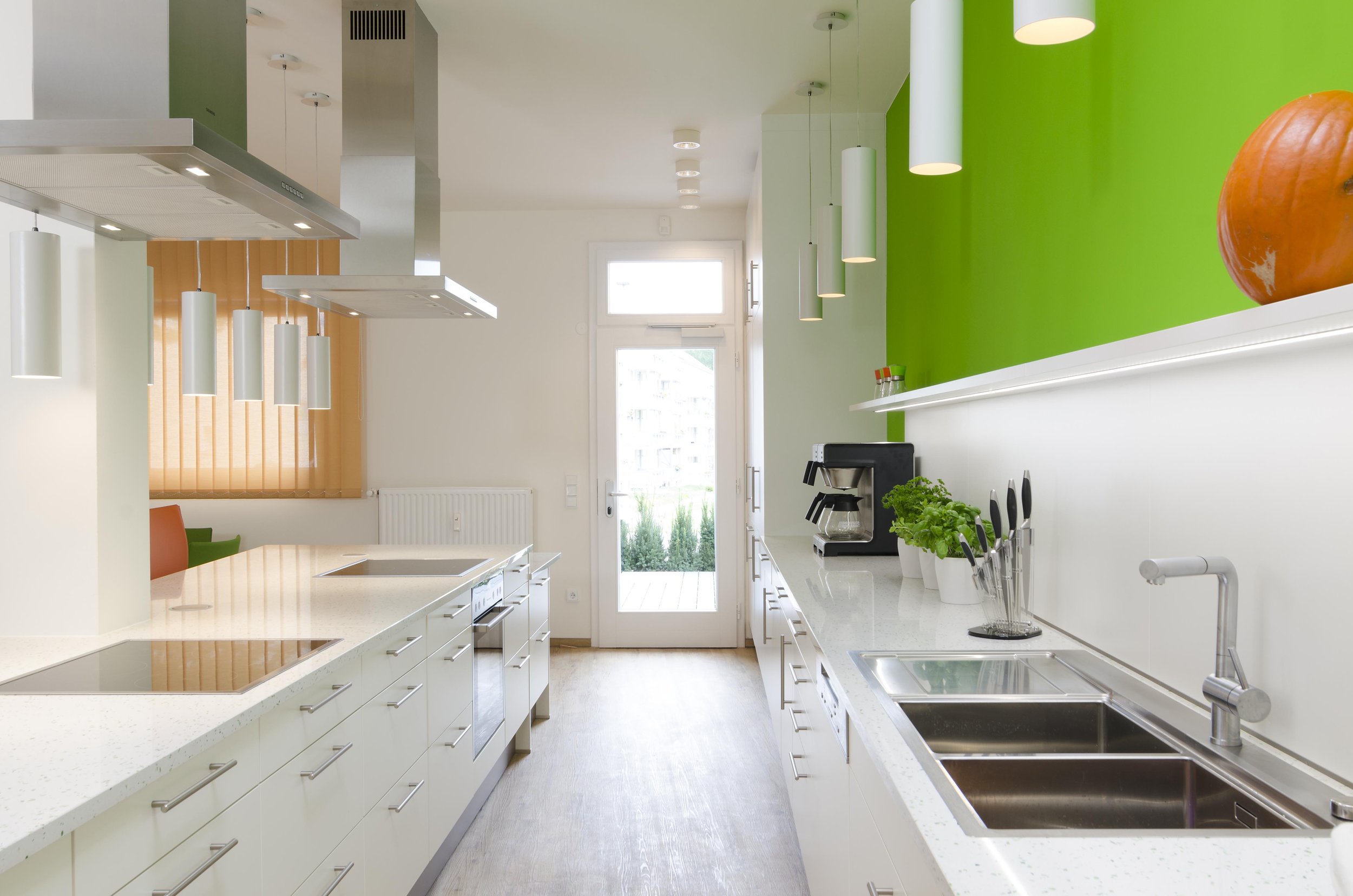 Moderne Küche mit weißen Schränken, grüner Akzentwand, Holzboden, Kochinsel, Herd, Backofen, Kaffeemaschine, Küchenutensilien, Pflanzen und große Fenster