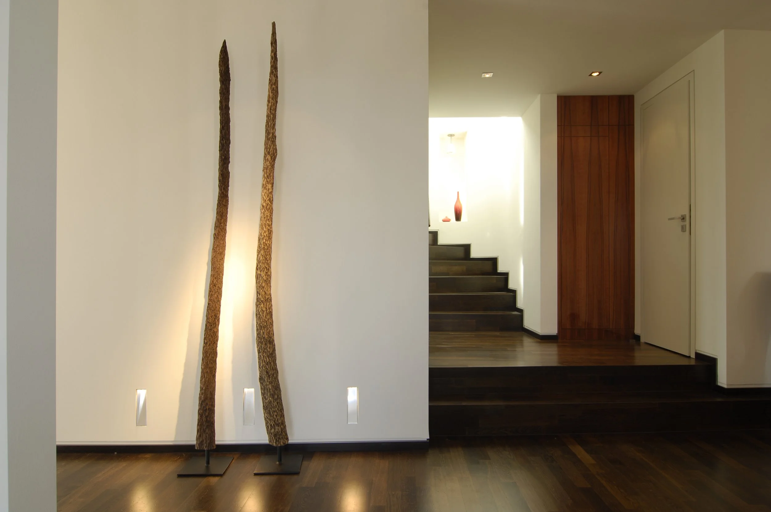 Innenraum eines modernen Hauses mit zwei Skulpturen aus Holz auf dem Boden, dunklem Laminatboden, weißen Wänden und einer Treppe mit dunklen Stufen im Hintergrund.