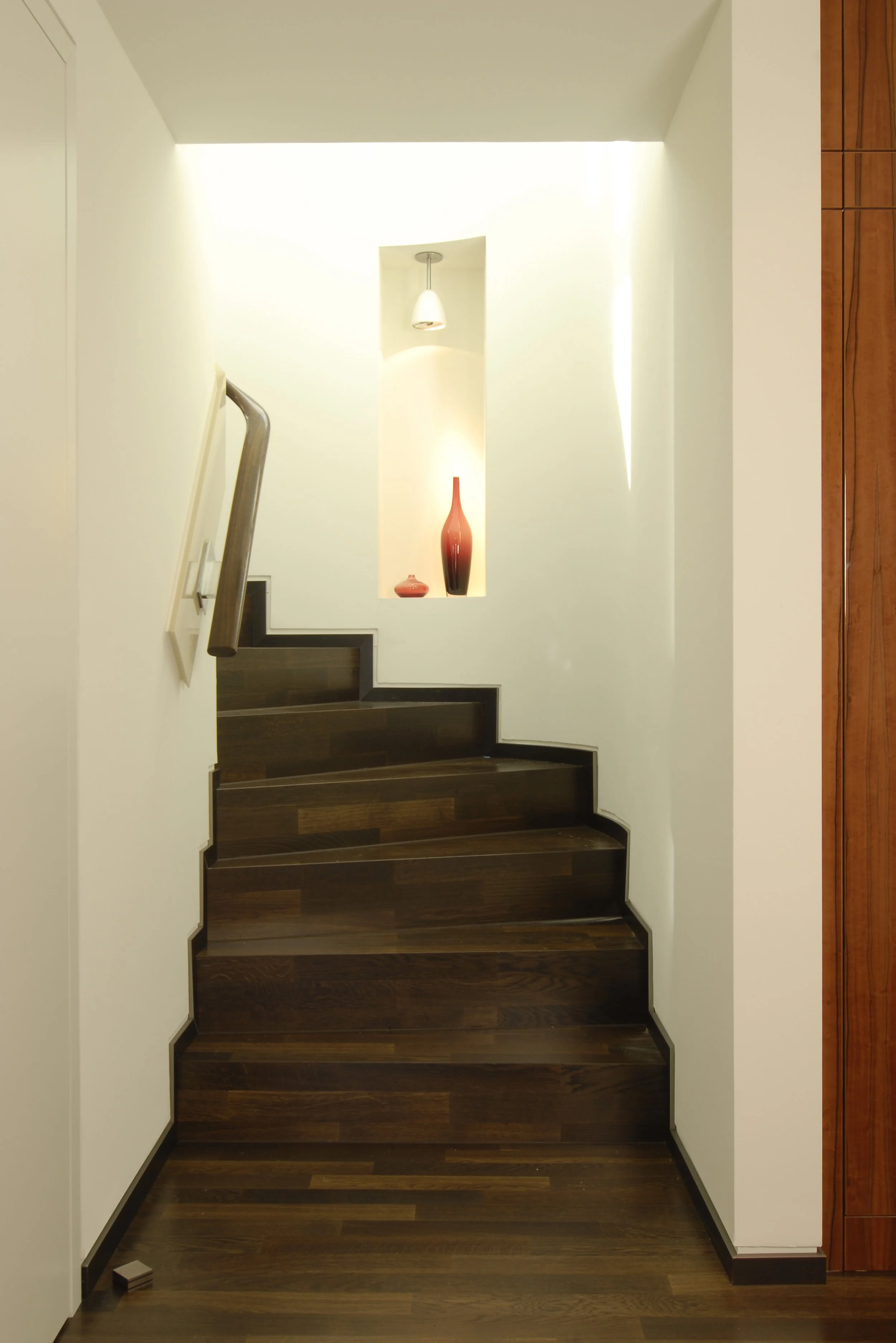 Treppenhaus mit dunklen Holztreppen, weißen Wänden und einer kleinen Nische mit Lampen und roten Dekorvasen