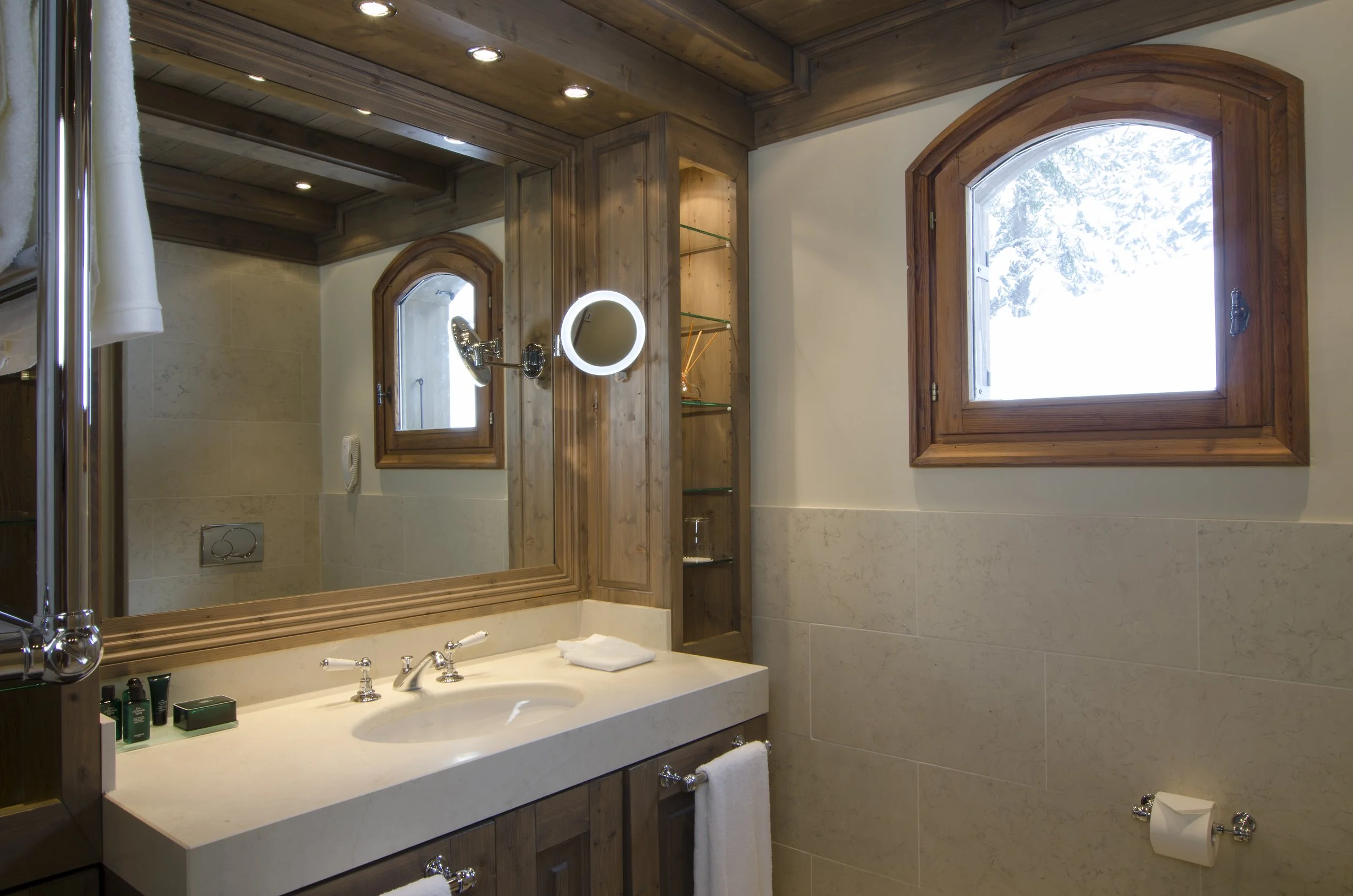 Badezimmer mit einem Holzrahmen Spiegel, einer Glasregalwand, zwei Fenster mit Holzkasten, Handtuchhalter und einem Waschtisch mit Seife, Shampoo und einem Handtuch.