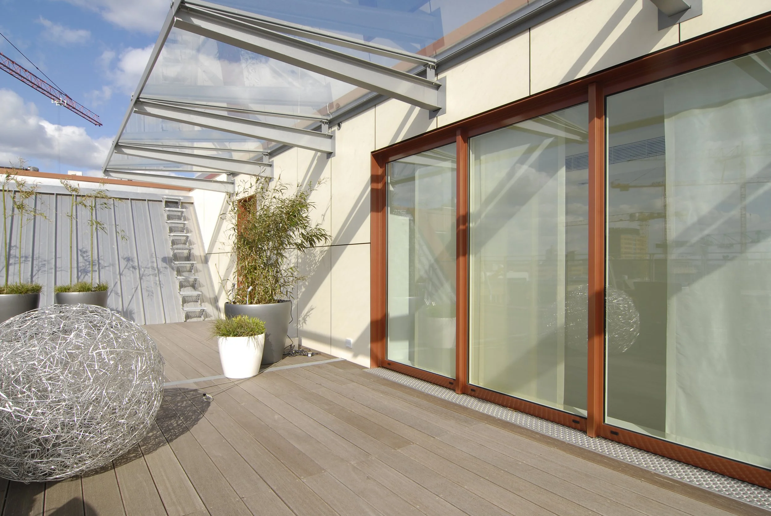 Blick auf eine moderne Terrasse mit Holzdielen, großen Pflanzkübeln mit Pflanzen, großen Glas-Schiebefenstern mit roten Rahmen, einem Sonnenschutz aus Glas und einem Himmel mit Wolken.