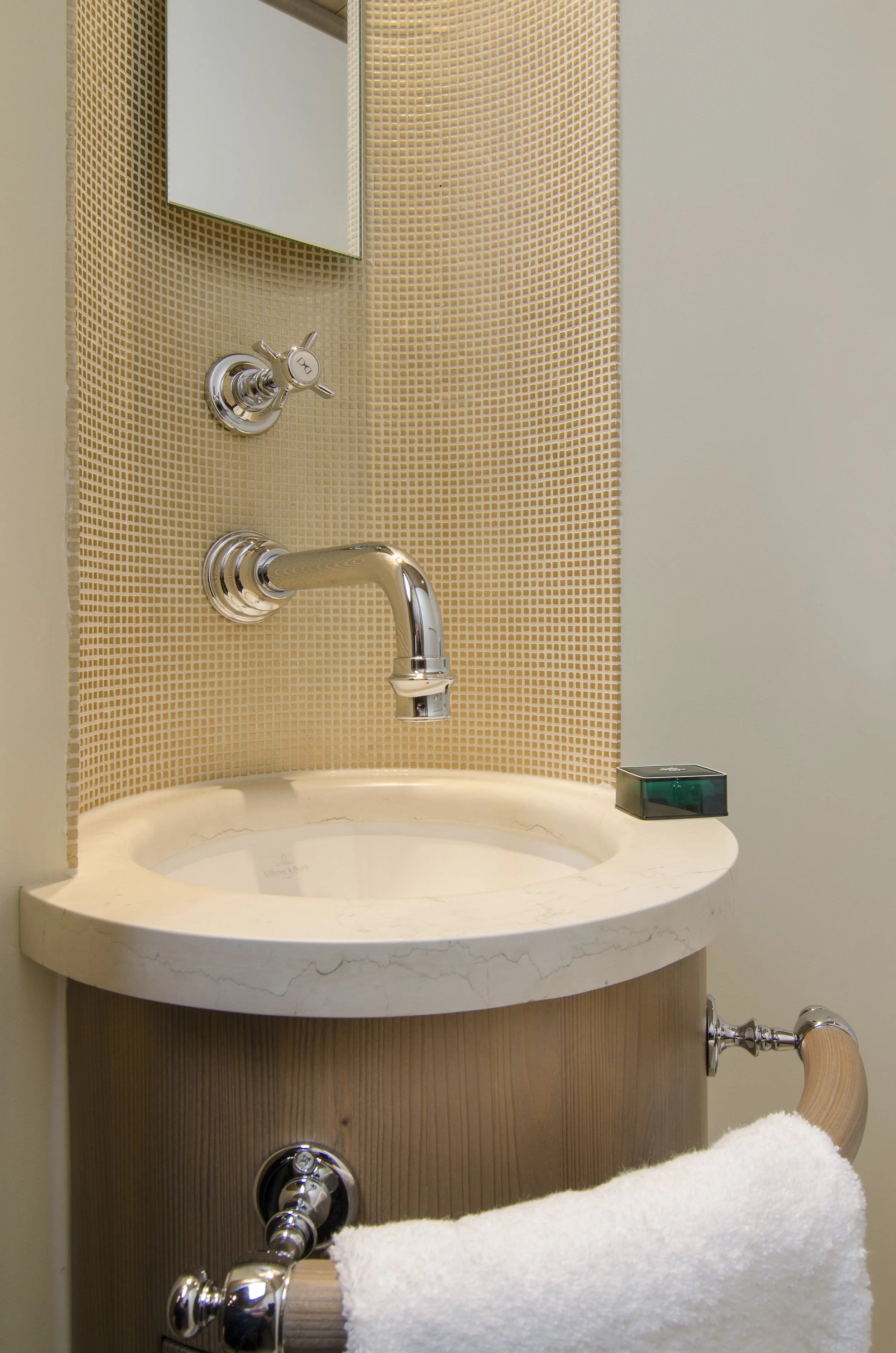 Ein Waschbecken mit Chrom-Armatür, einem Spiegel, einer kleinen schwarzen Box auf der rechten Seite, einer weißen Handtuchrolle auf einer Holzhalterung, vor einer beige Fliesenwand.