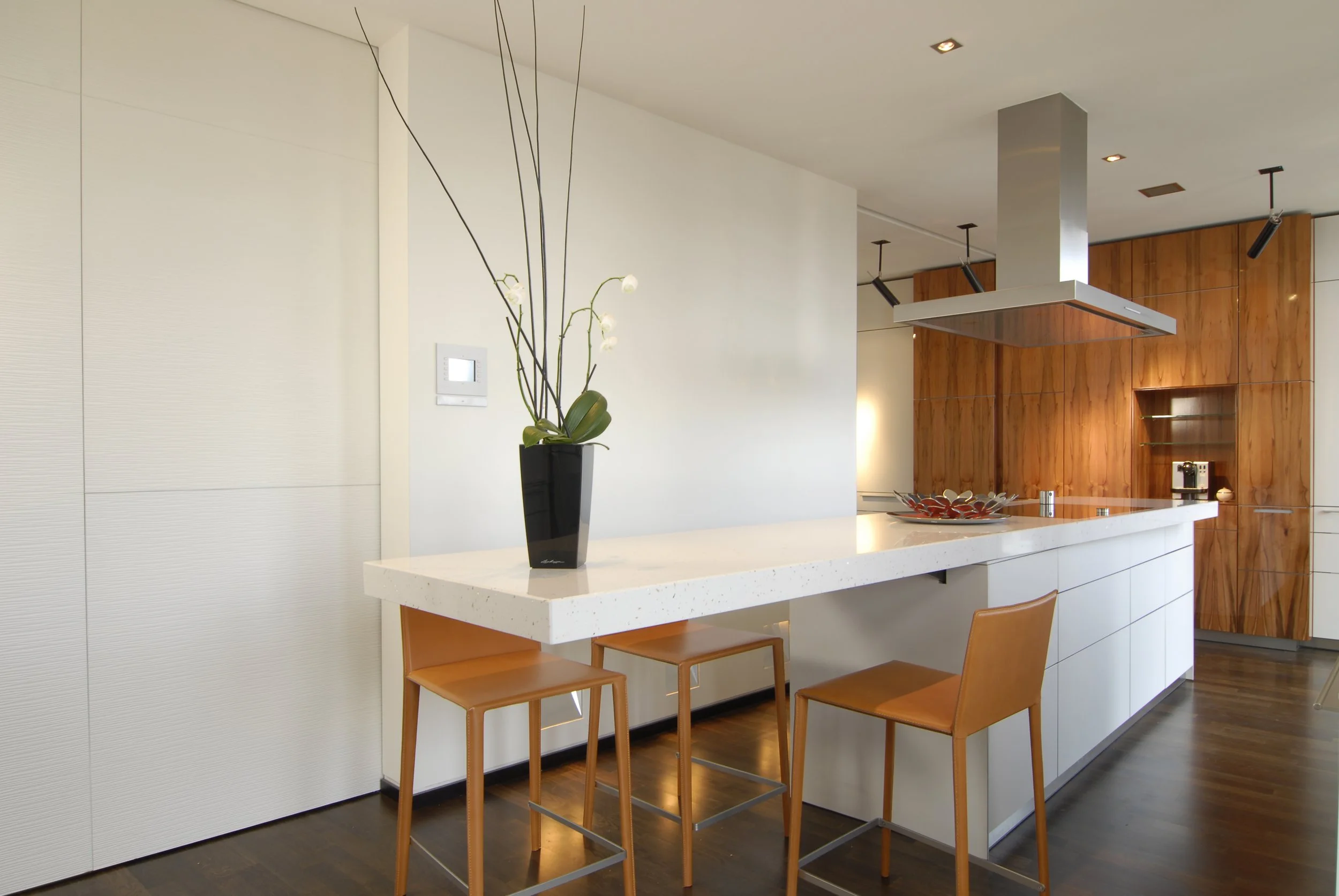 Moderne Küche mit weißen Schränken, Holz-Elementen und einem weißen Frühstückstresen mit zwei braunen Stühlen, einer schwarzen Pflanze auf dem Tresen, befriedeter Holzfußboden, elegante Beleuchtung