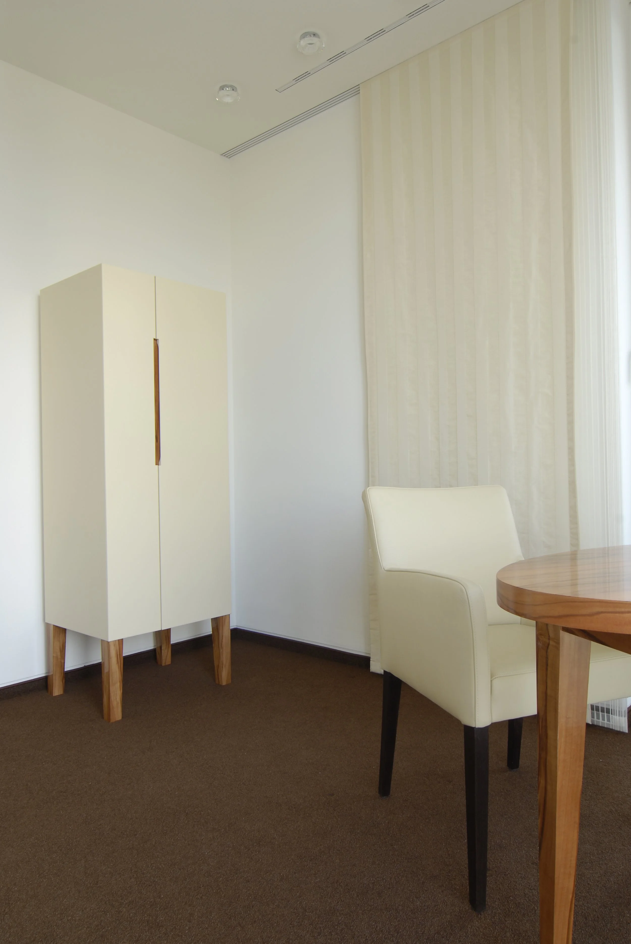 Ein stilvoll eingerichteter Raum mit weißem Kleiderschrank, einem weißen Sessel und einem runden Holztisch, mit weißen Gardinen vor einem Fenster.