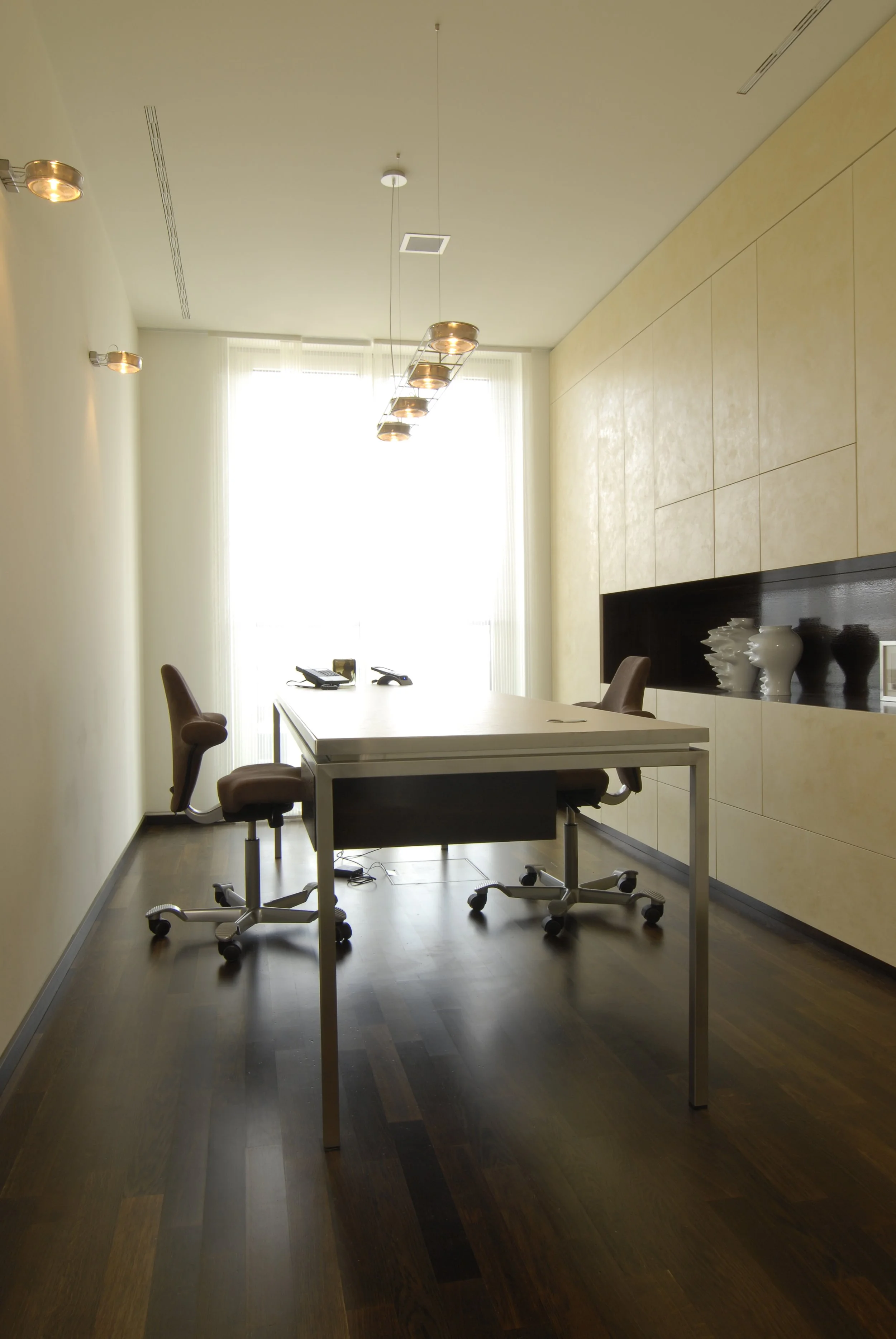 Ein modernes Büro mit einem weißen Tisch, drei braunen Bürostühlen, dunklem Holzboden, und einer Wand mit Ablagefach in schwarzen und cremefarbenen Tönen. Helle Gardinen lassen viel Licht herein.