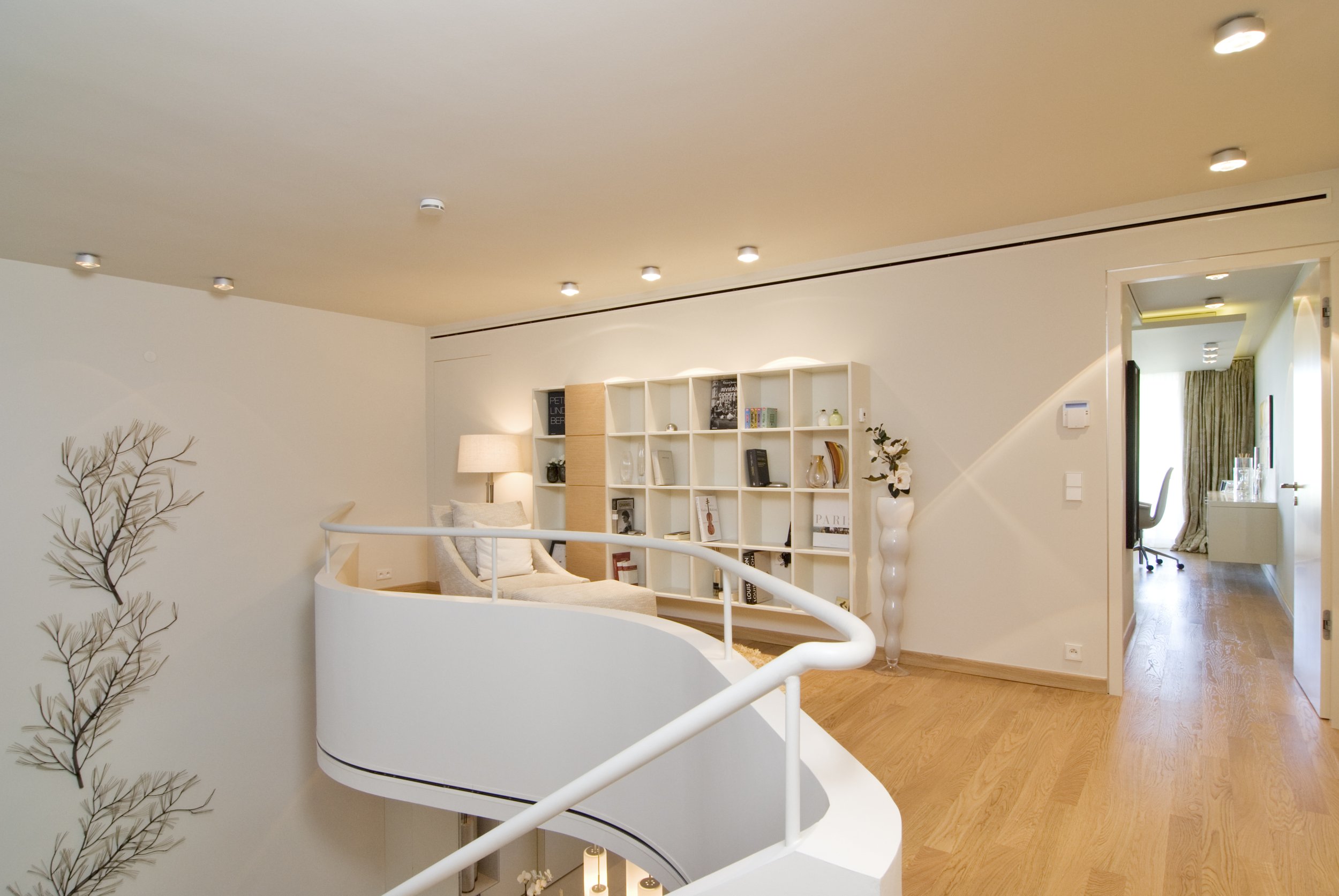 Helles, modernes Wohnzimmer mit weißem Bücherregal, Lesesessel und Dekorationsobjekten auf Holzboden, neben einer Treppe und einem Flur mit Schreibtisch.