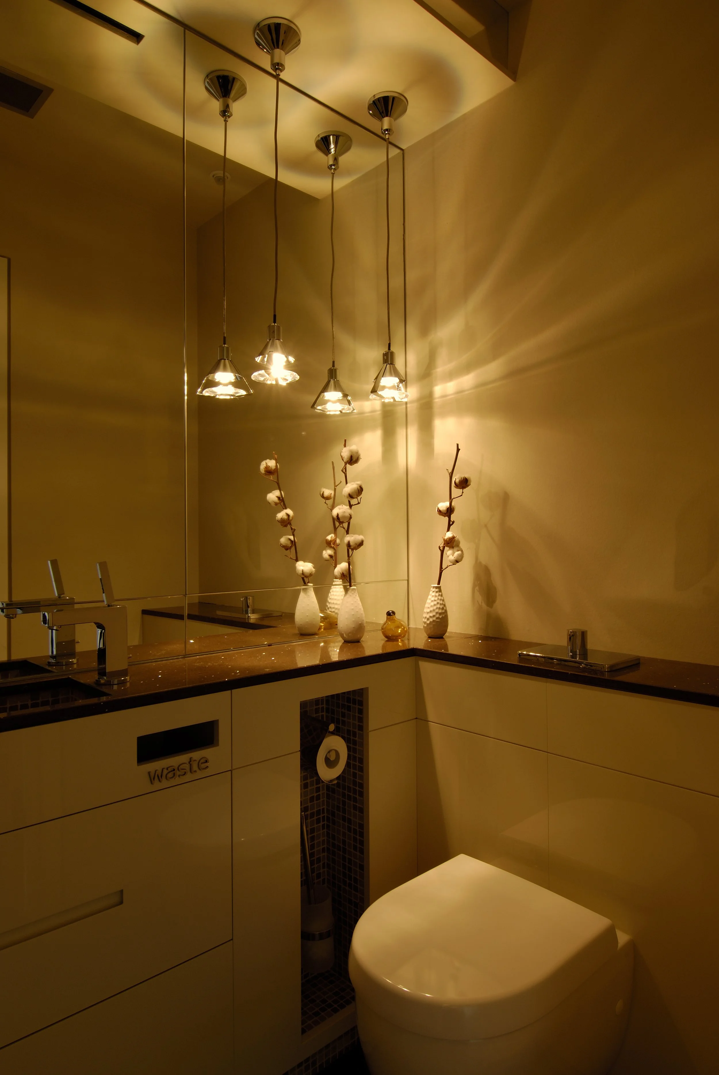 Moderner Badezimmerbereich mit großen Spiegel, decorativen Vasen mit Baumwollzweigen, angeleuchtete Deckenleuchten, WC, und verschiedenen Abteilen.
