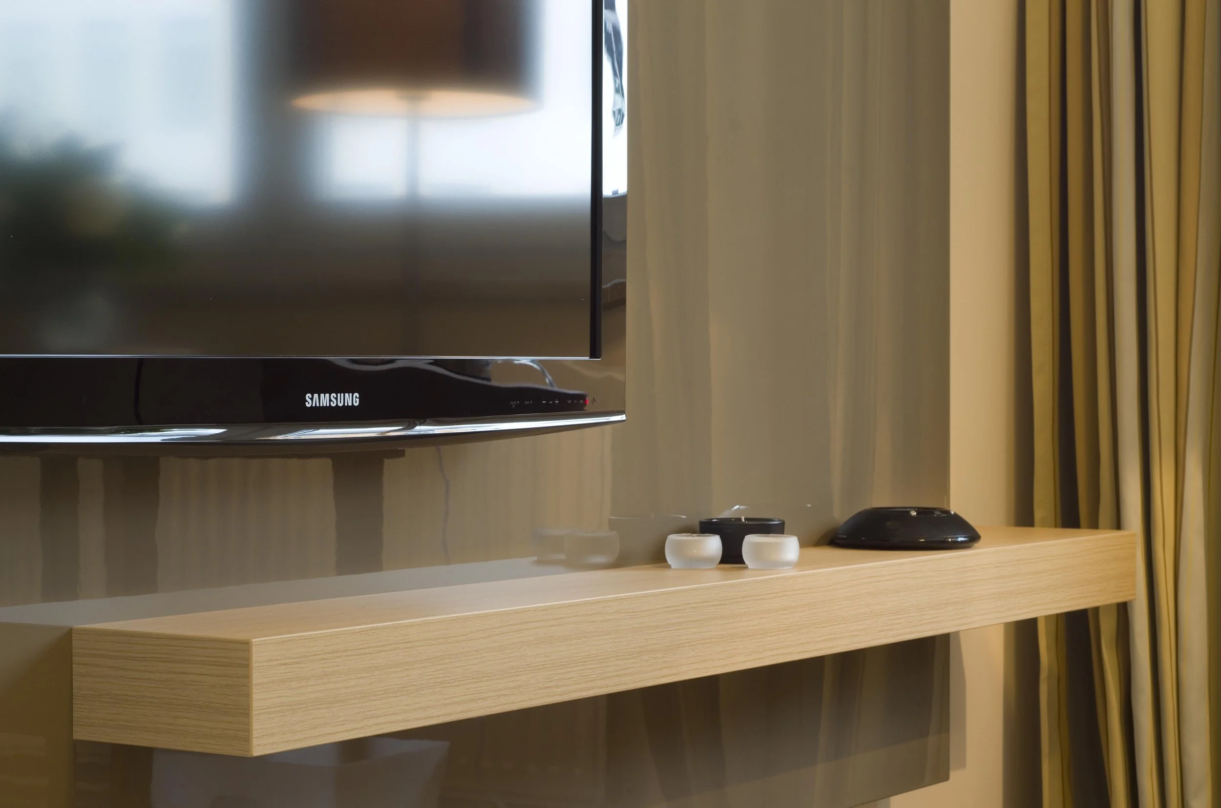 Ein Fernseher auf einem Holzregal mit kleinen Dekogefäßen und einem schwarzen elektronischen Gerät auf der rechten Seite, neben einer Gardine.
