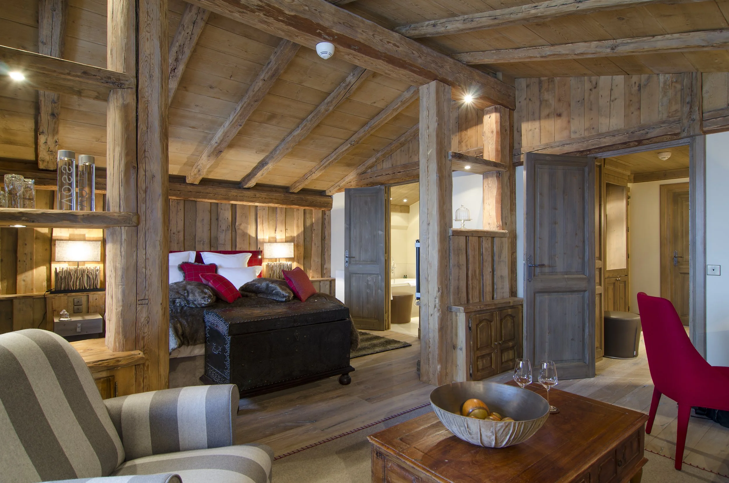 Ein gemütliches Schlafzimmer mit Holzverkleidung, einem Bett mit weißen und roten Kissen, einer Truhe am Fußende, seitlichen Nachttischen mit Lampen, ein erdiges Farbschema, und ein Wohnzimmerbereich mit einem grauen und weißen gestreiften Sessel, ei