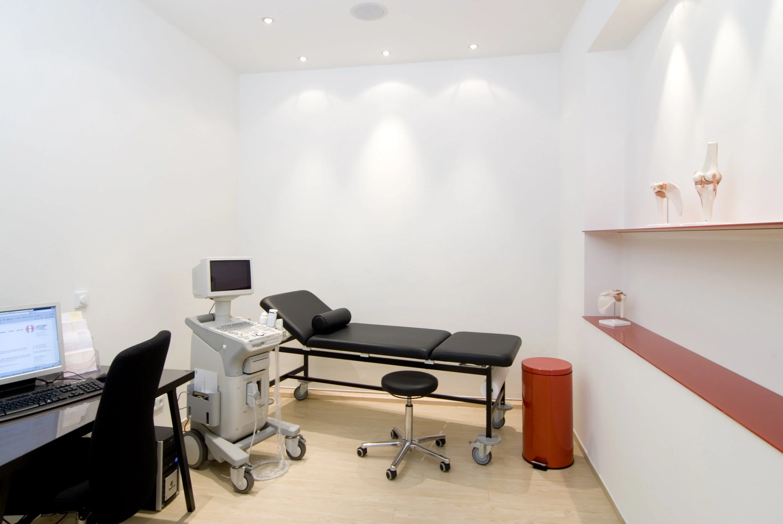 Ein medizinisches Behandlungszimmer mit Untersuchungsliege, Computer, medizinischen Geräten, Stuhl und Dekorationsstücken an der Wand.