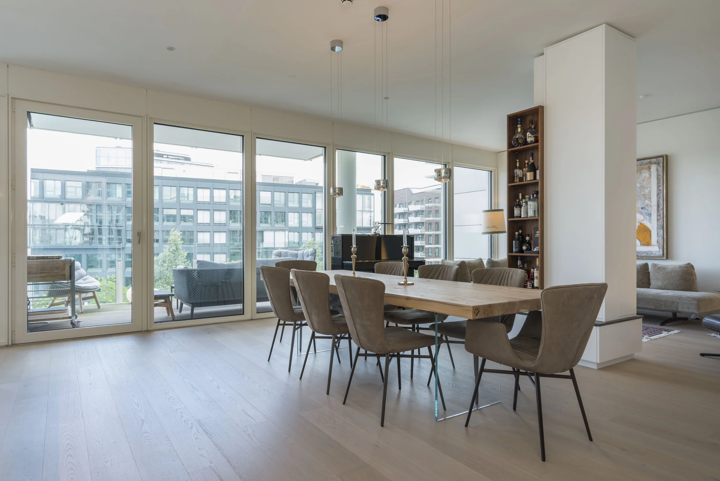Helles, modernes Wohnzimmer mit großen Fenstern, Esstisch mit neun Stühlen, offener Zugang zur Terrasse, Wohnzimmer mit Sofa, Kunstwerk an der Wand, Bücherregal mit Spirituosen, Holzboden.